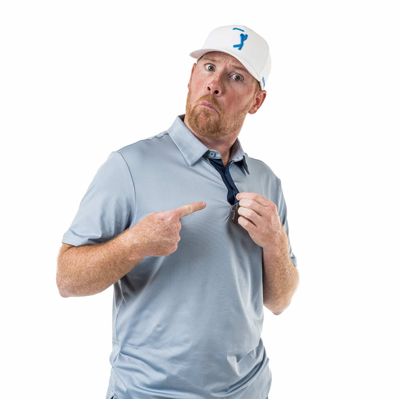 A man with a reddish beard in a light gray polo and white cap looks surprised, pointing at his shirt collar and tugging it, standing against a plain white background.