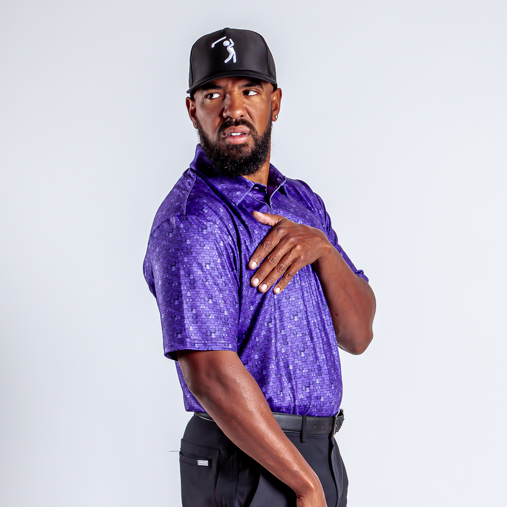 A man in a purple patterned polo shirt and black cap touches his left shoulder and looks to his left with a serious expression, standing against a plain white background.