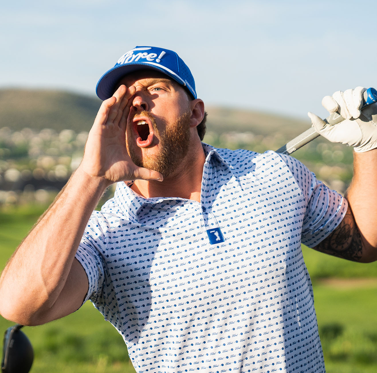 A bearded man in a blue cap and patterned golf shirt shouts with his hand at his mouth while holding a golf club, standing outdoors on a sunny golf course.