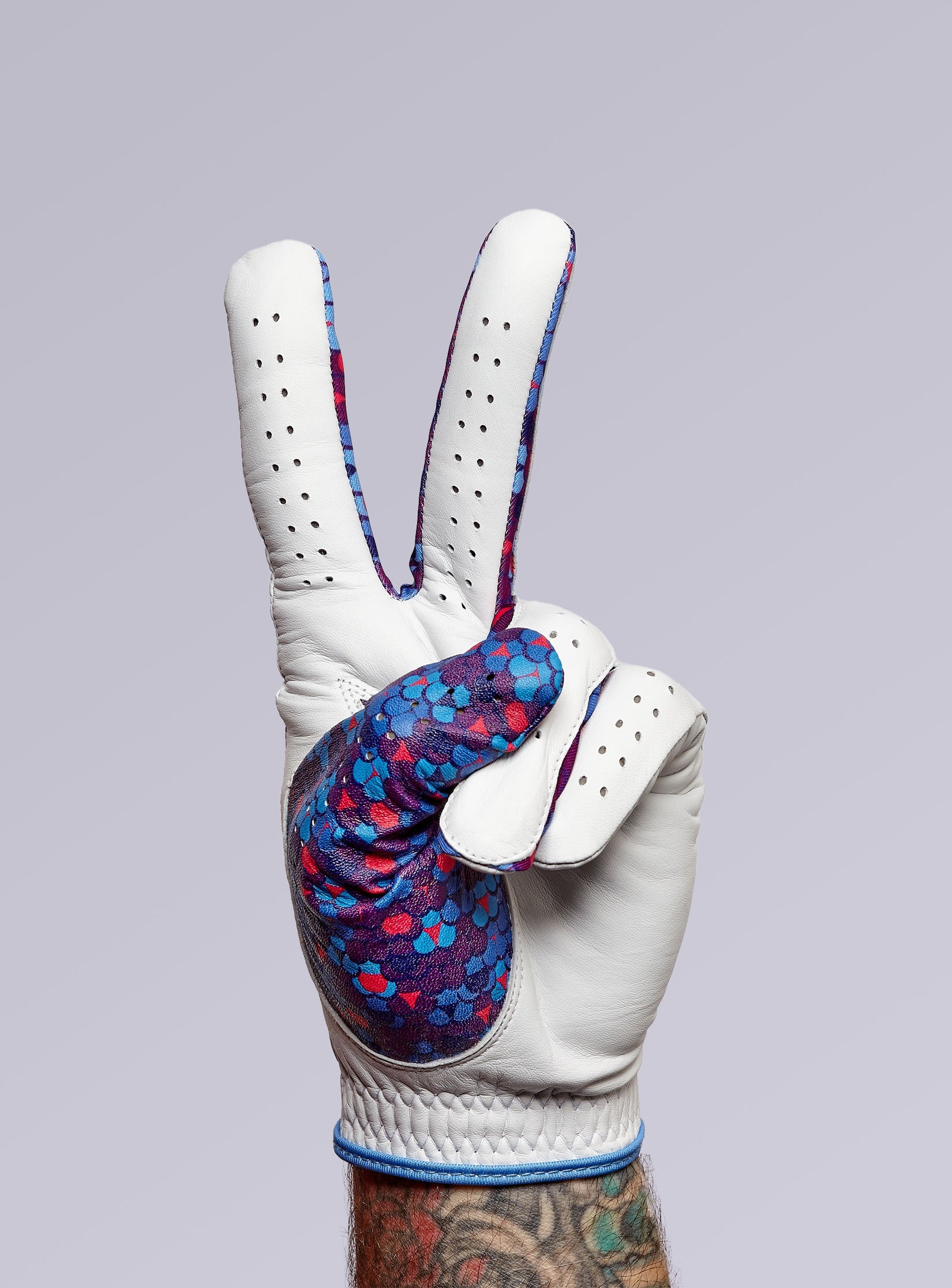 A hand wearing a white glove with colorful blue and red patterns on the palm shows a peace sign against a light purple background. The wrist has visible tattoos.