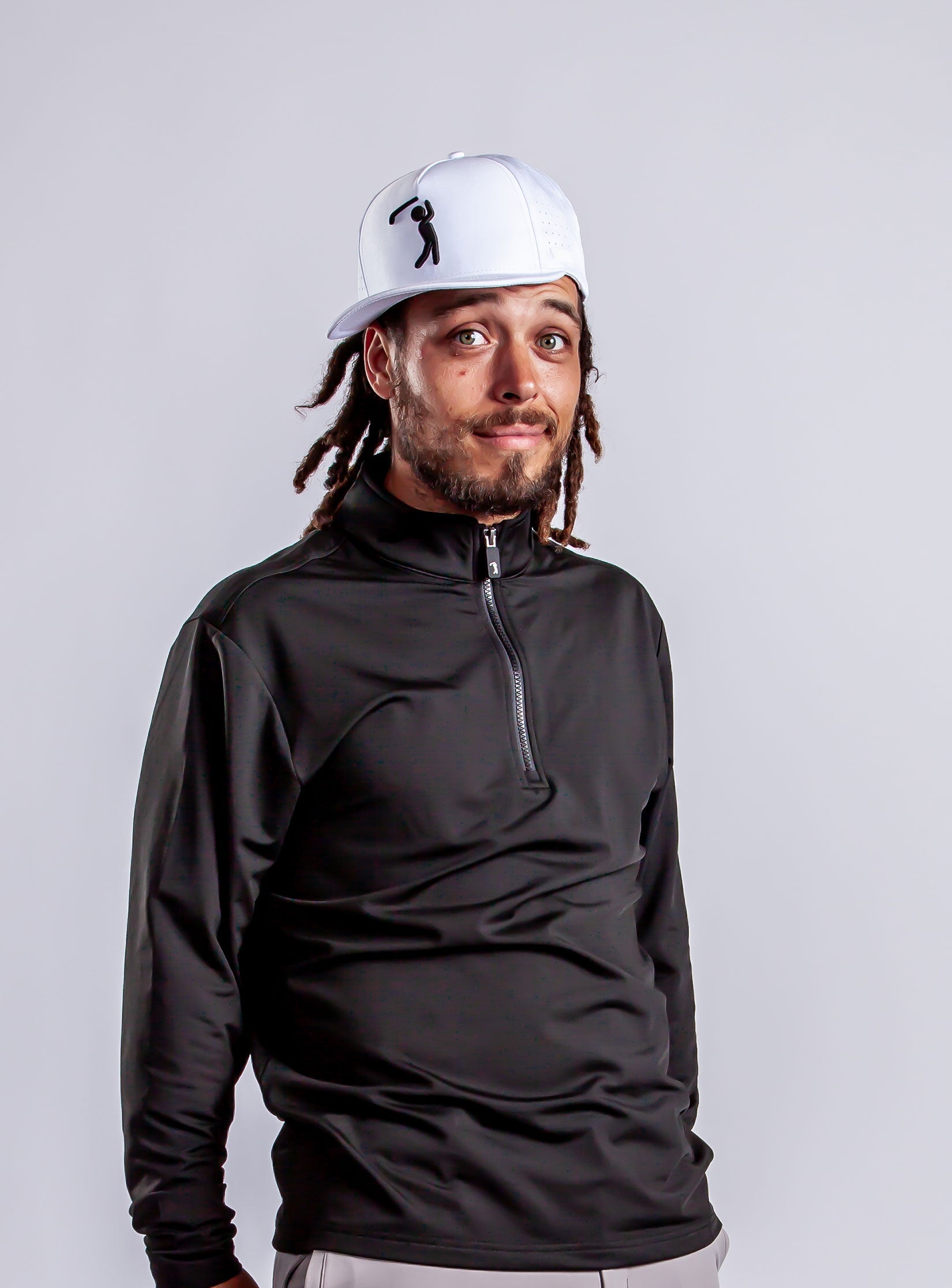 A man with long dreadlocks wearing a white cap sideways and a black zip-up shirt stands against a plain light gray background, looking at the camera with a slight smile and raised eyebrows.