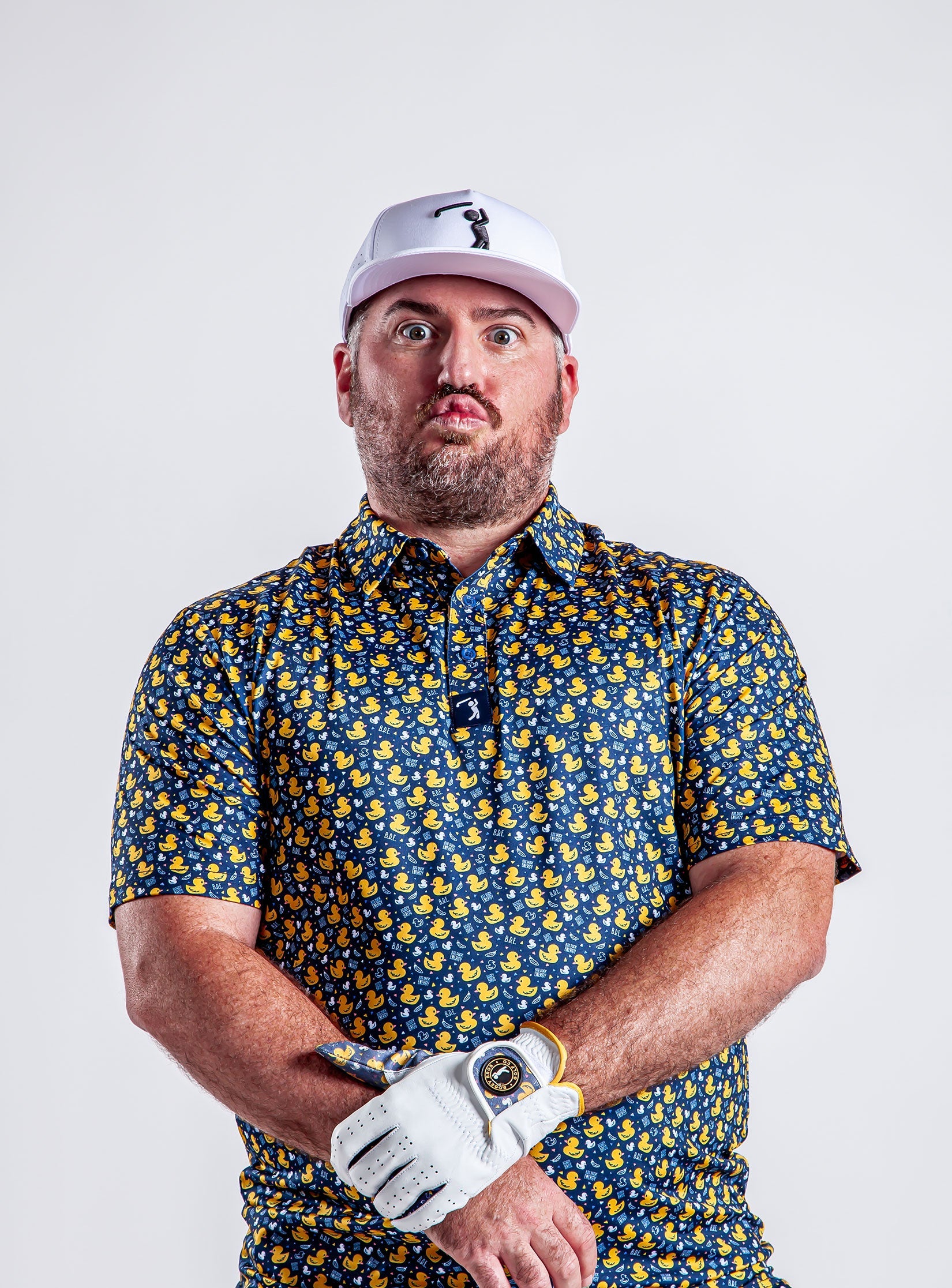 A man in a duck-print shirt and white cap makes a funny, surprised face. He wears a white golf glove and stands against a plain light background.