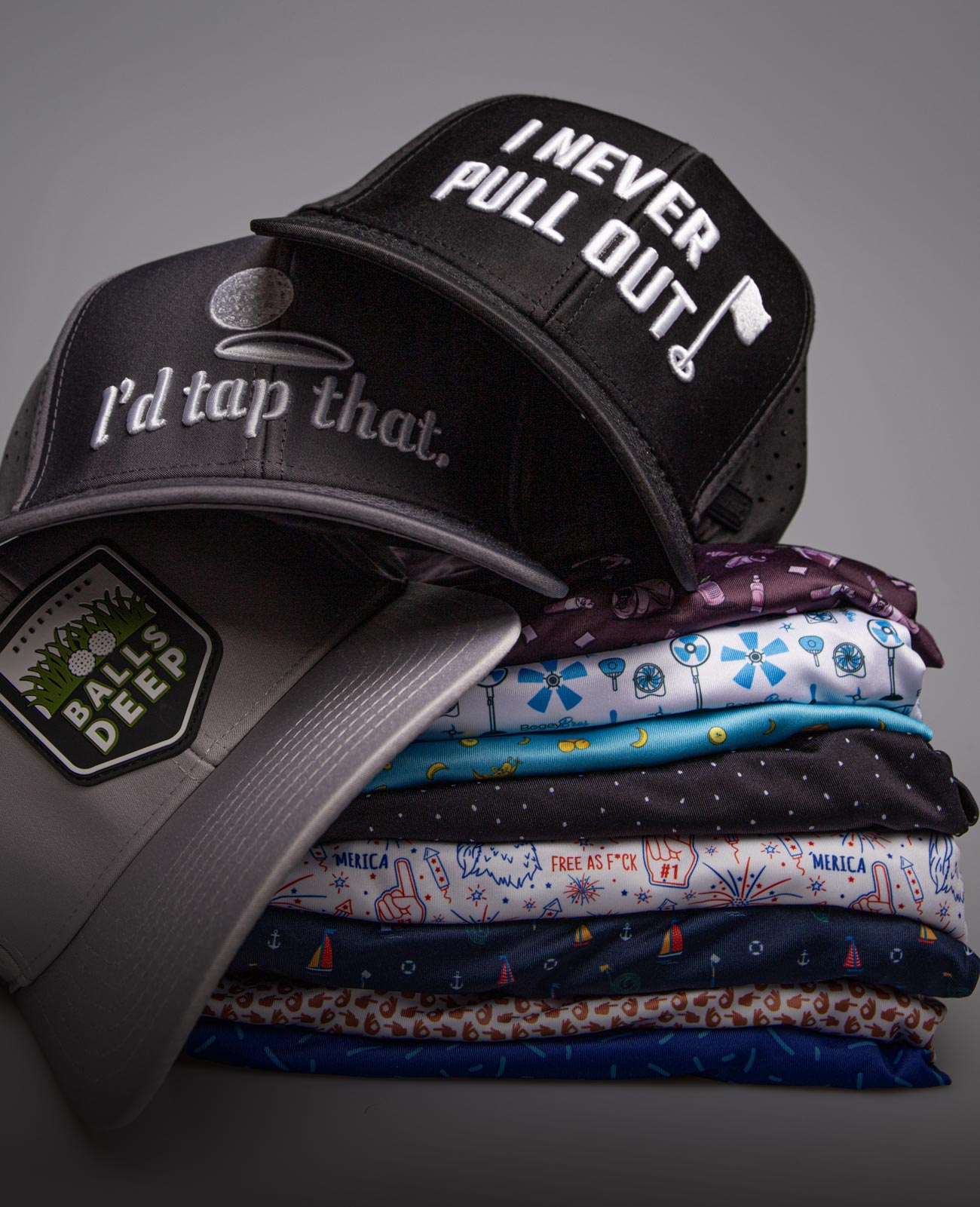 A stack of colorful, patterned shirts topped with three hats. One hat reads “I NEVER PULL OUT” with a golf flag, another says “I’d tap that.” with a golf hole, and the third has a patch reading “BALLS DEEP.”.