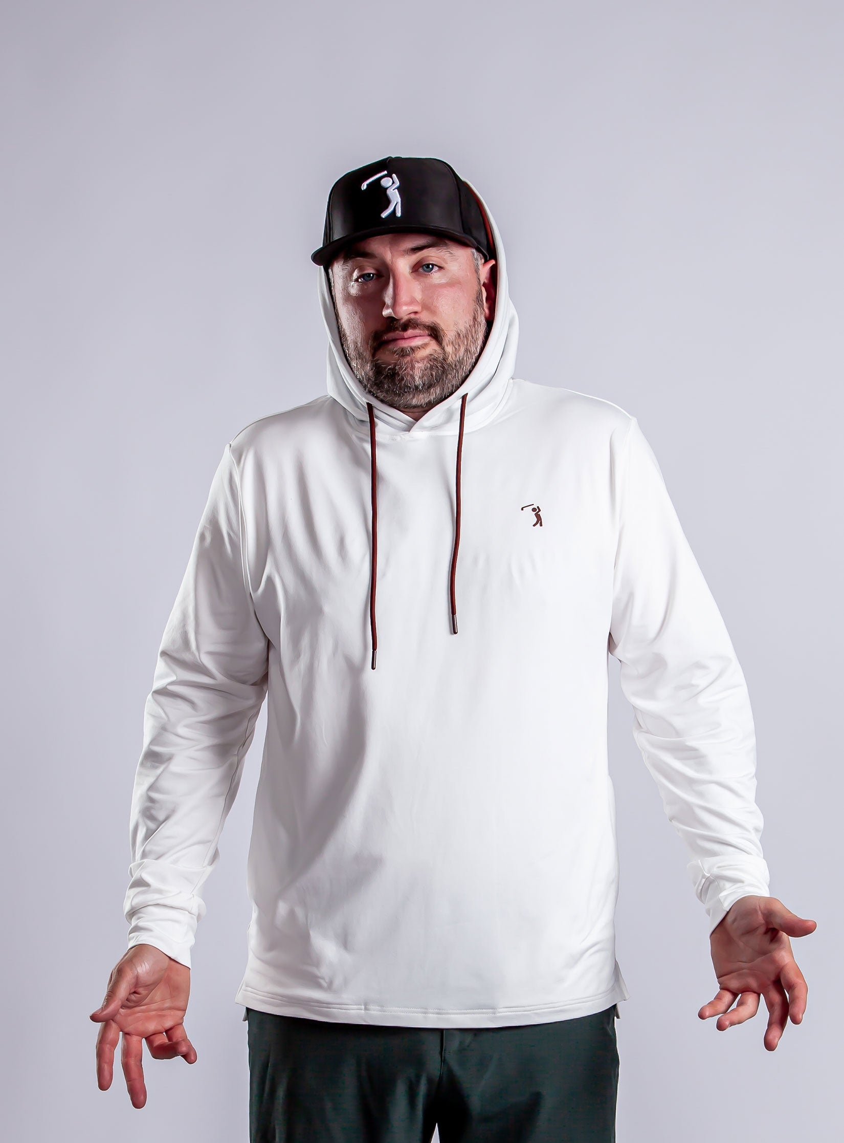 A bearded man in a white hooded sweatshirt and black cap stands against a plain background, hands out and palms up, with a neutral expression. Both his hoodie and hat have a small golfer logo.