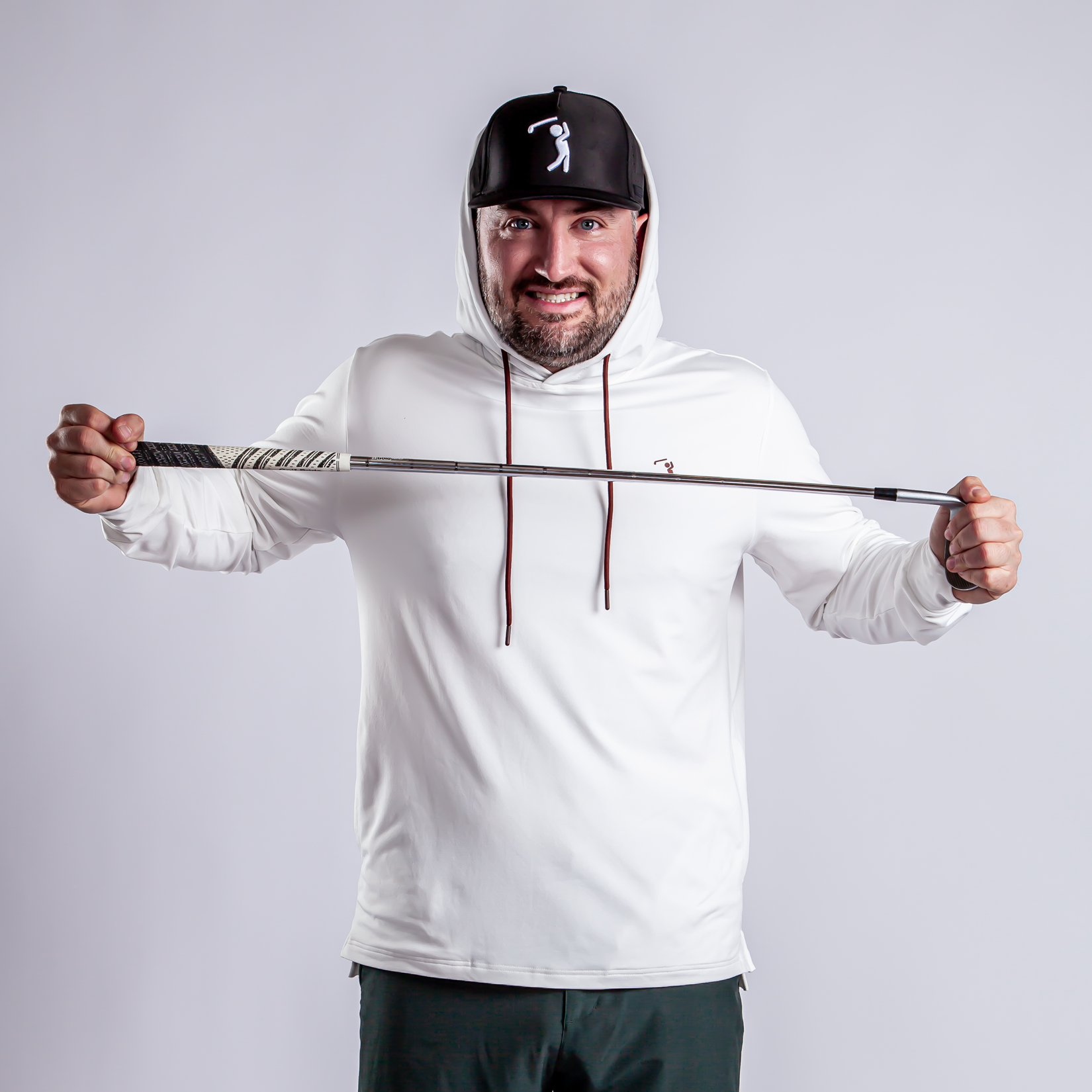A smiling man in a white hoodie and black cap holds a golf club horizontally with both hands, facing the camera against a plain light gray background.
