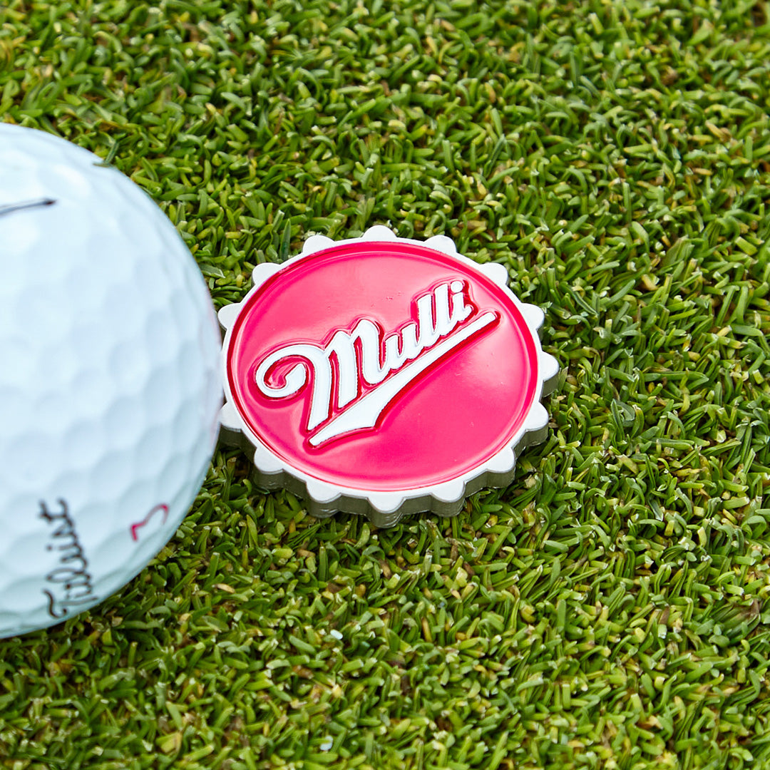 A white golf ball sits on green grass next to a red and white bottle cap-shaped marker with the word Mull written in script across the top.