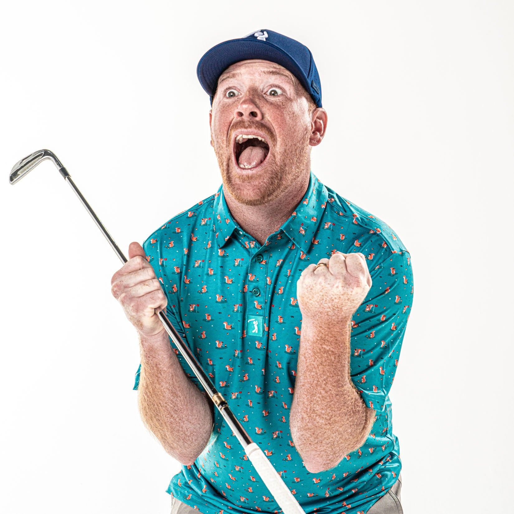 A red-haired man in a blue patterned shirt and blue cap clenches his fists and shouts excitedly while holding a golf club, expressing intense joy against a white background.