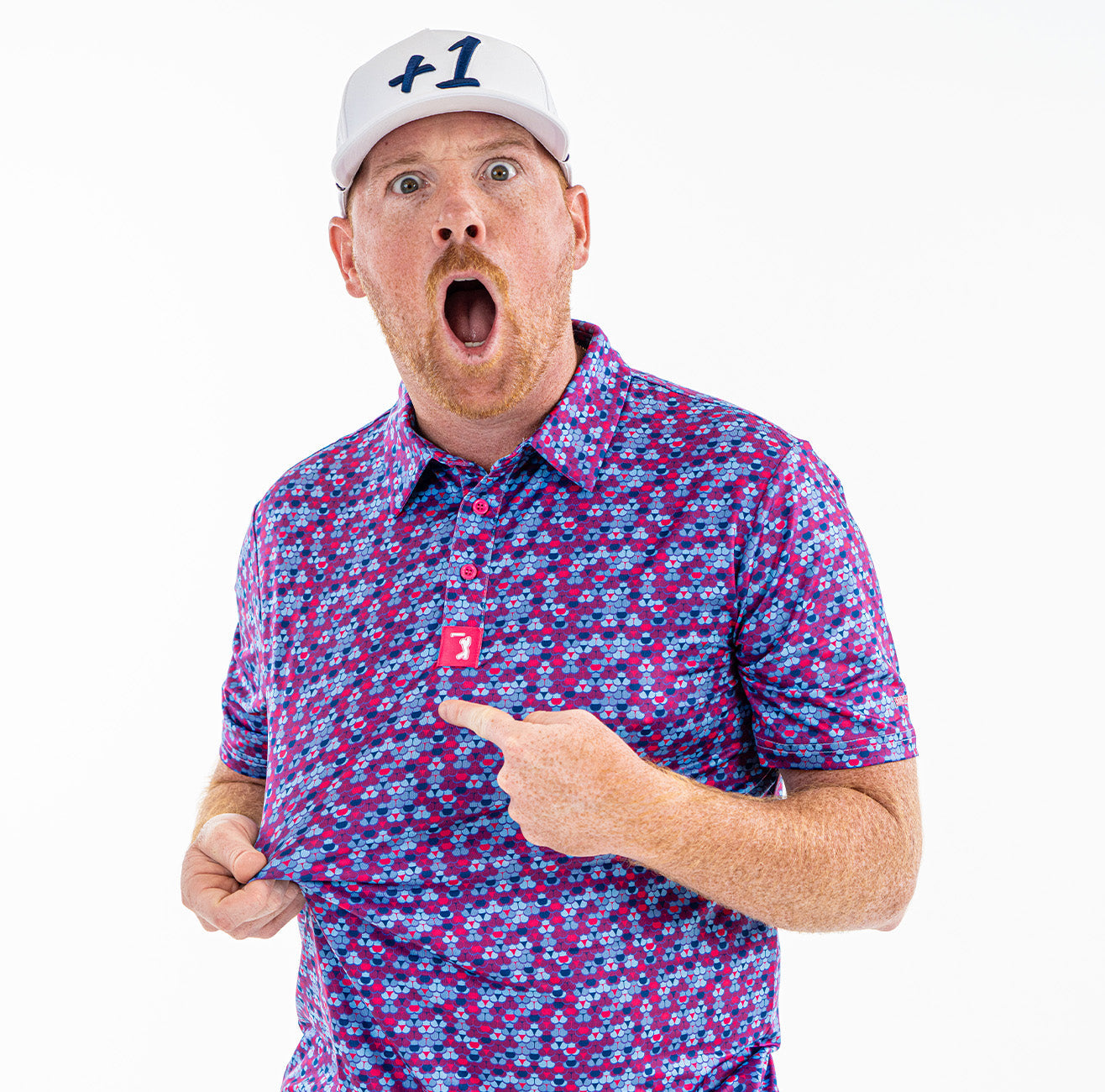 A surprised man in a white “+1” cap and colorful purple patterned shirt holds out his shirt and points to a red tag, mouth open wide, standing against a white background.