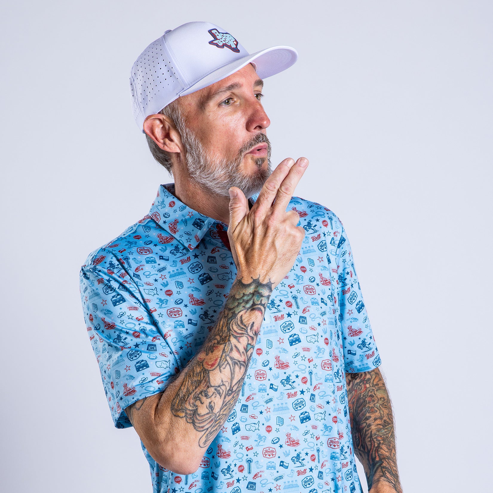 A man with a gray beard and tattooed arms wears a white cap and a blue patterned shirt. He faces left, holding his right hand up in a playful finger gun gesture, with a light gray background behind him.