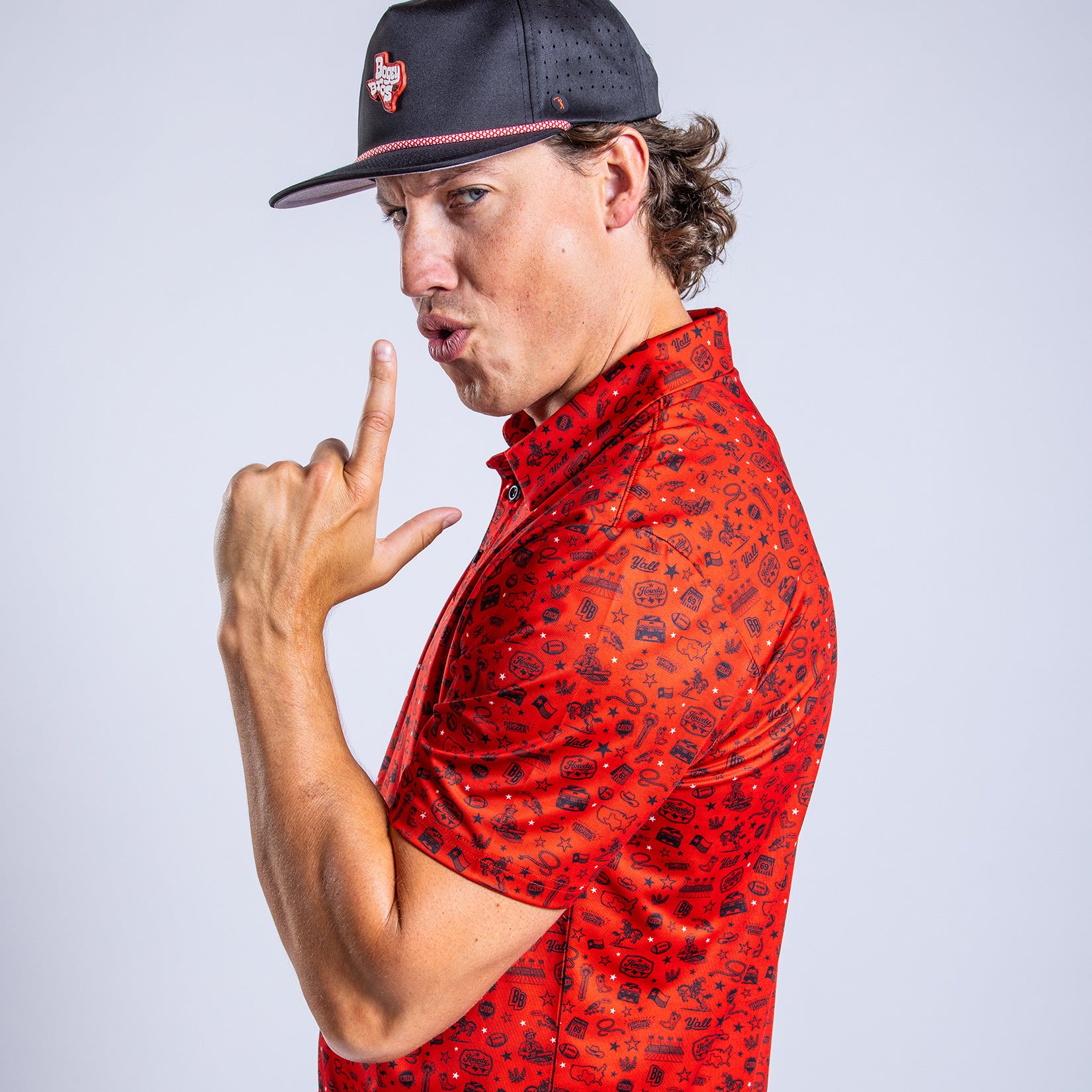 A man in a black hat and red patterned shirt stands sideways, making a playful finger gun gesture with his right hand while pursing his lips, set against a plain light gray background.