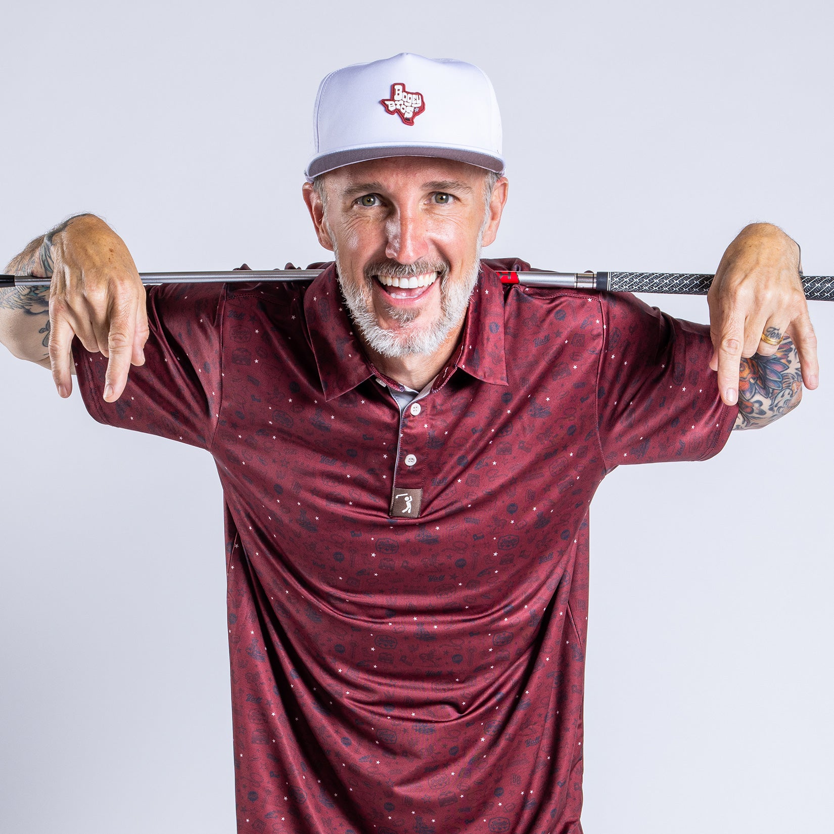 Smiling man with a short gray beard wears a maroon patterned polo shirt and a white baseball cap with a Texas emblem. He rests a golf club across his shoulders, holding it with both hands. Tattoos are visible on his arms. Gray background.