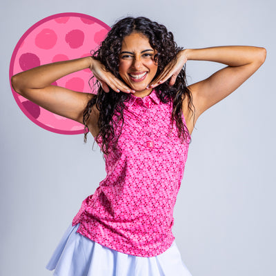 A smiling woman with curly hair poses with her hands framing her face, wearing the Dots - Pink - Women's Polo and a white skirt. A large pink circle with dark dots is behind her, set against a plain gray background.