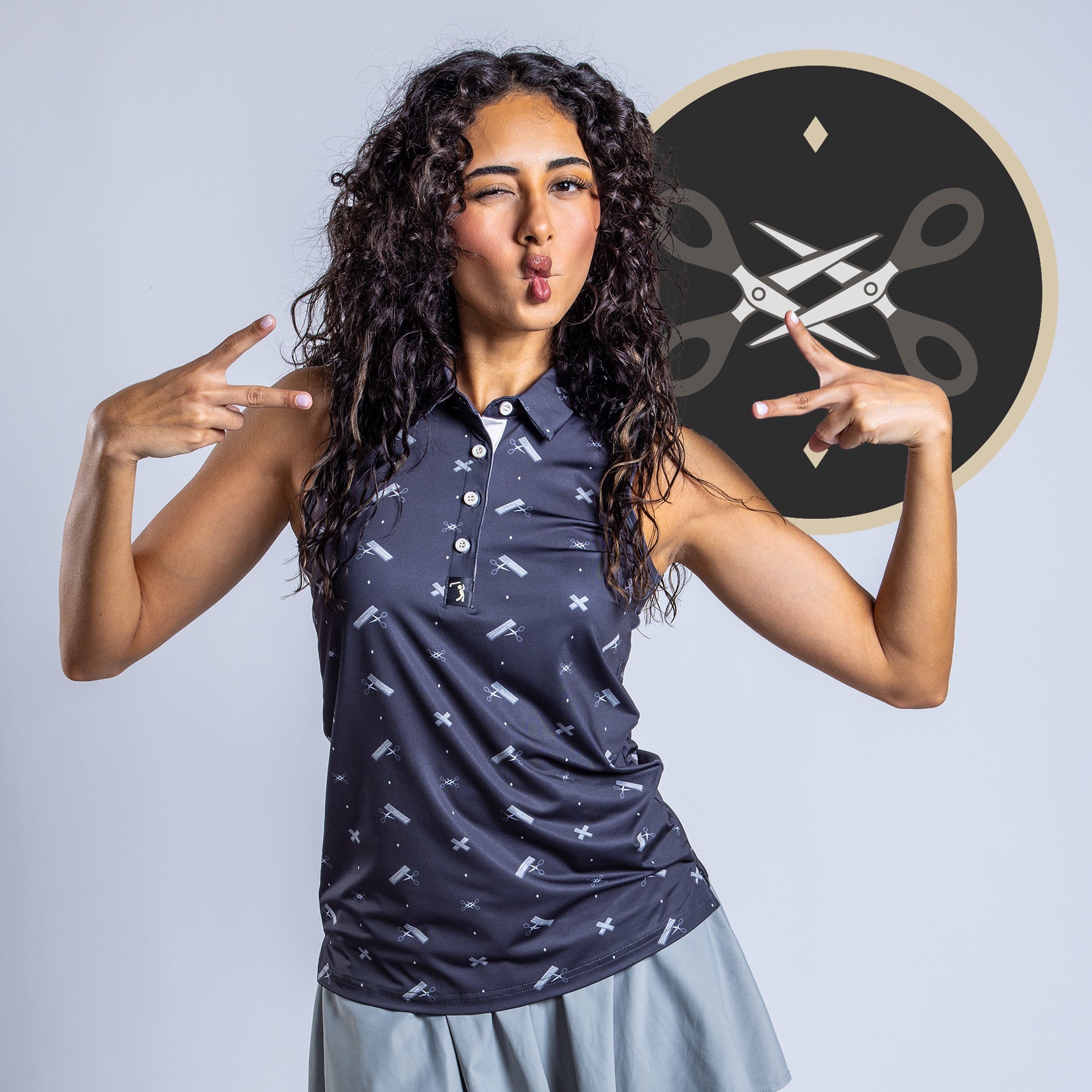 A curly-haired woman in a sleeveless, moisture-wicking navy Scissor Me Timbers - Women's Polo and light gray skirt makes playful peace signs. Behind her is a circular graphic featuring scissors, a ruler, and pencil on a dark background.