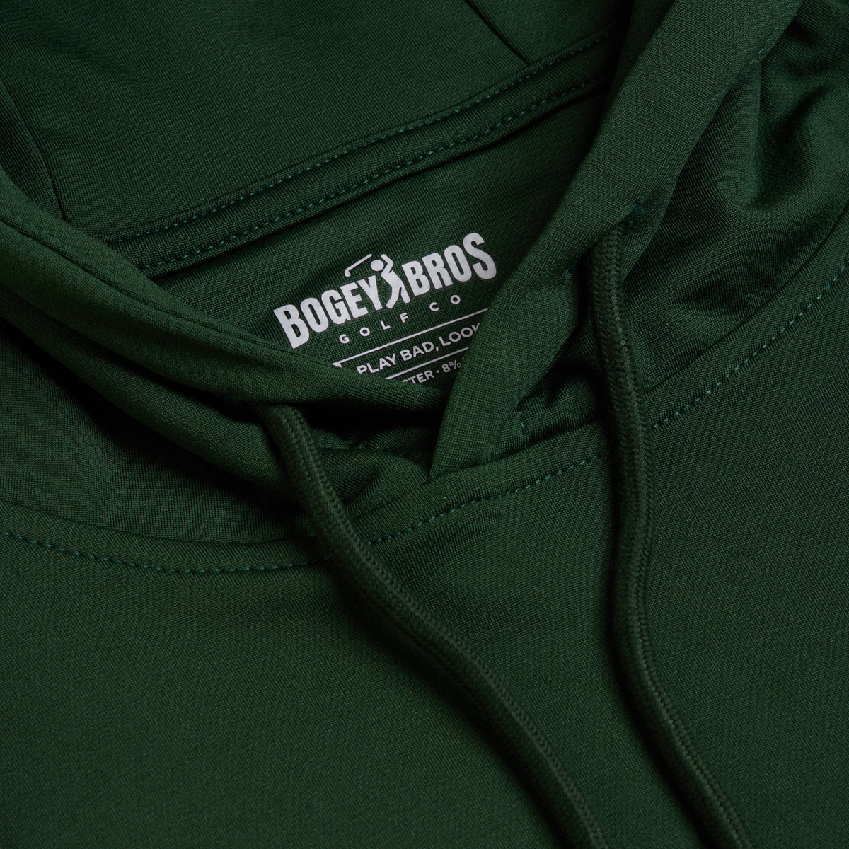 A close-up of the Forest Green - Hoodie highlights the hood’s drawstrings and a white inner label with the text “Bogey Bros Golf Co. PLAY BAD, LOOK GOOD.” The image clearly captures the textured, stretchy fabric and detailed stitching—reminiscent of moisture-wicking athletic shorts—focusing on the upper chest and neck area.
