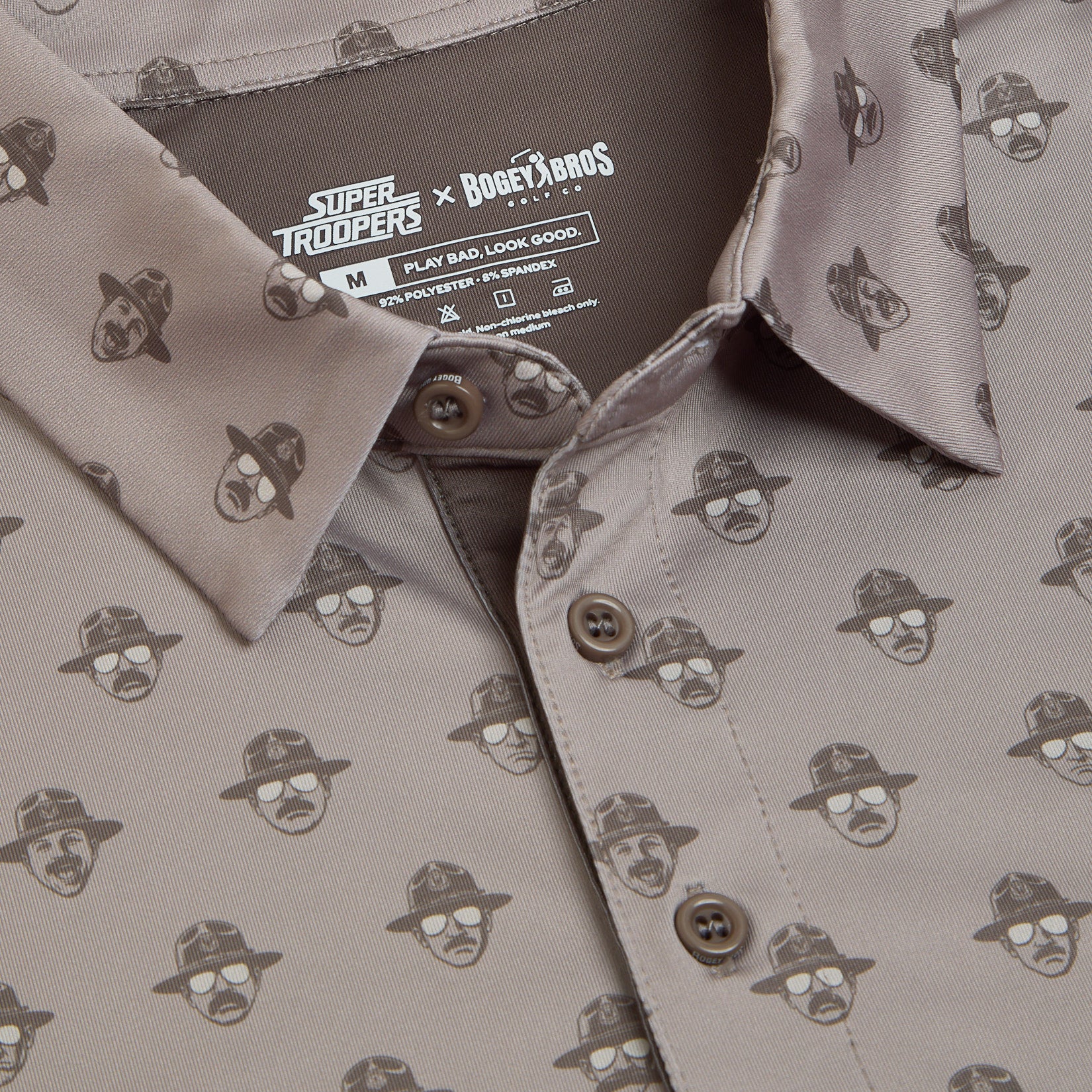 Close-up of a beige button-up shirt featuring a repeating pattern of cartoon faces wearing sunglasses and ranger hats. The collar and shirt tag show collaborations: Super Troopers x Bogey Bros. Care instructions and size M are visible inside the collar.