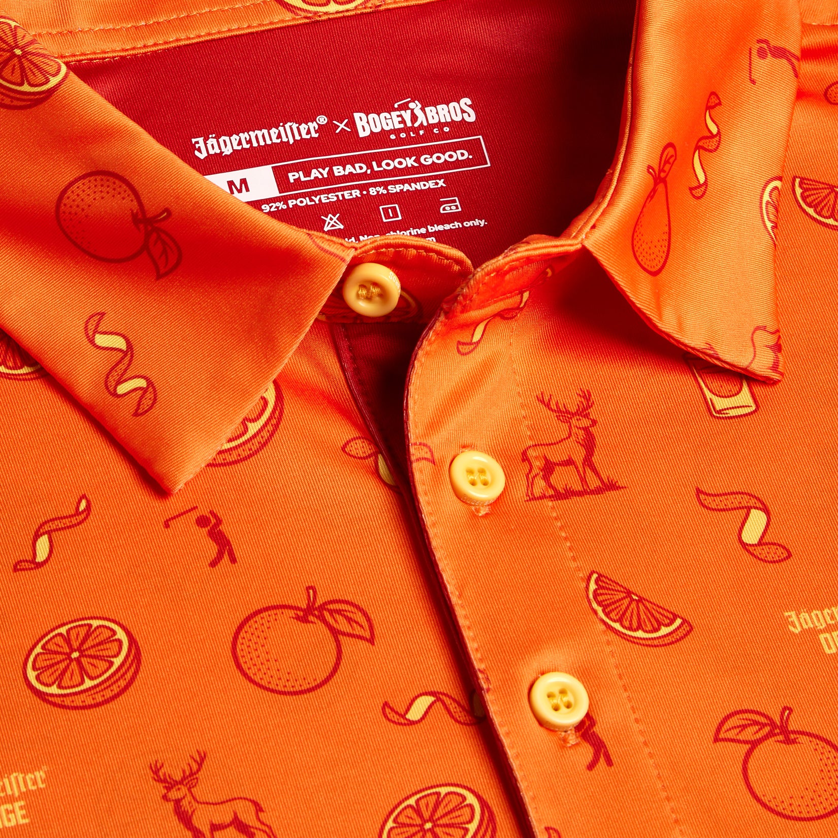 Close-up of the Main Squeeze - Polo in orange with playful red line art of oranges, deer, citrus slices, bottles, and ribbons. Features an athletic fit, moisture-wicking fabric, and a Jägermeister x BOGEY BROS collar label. Play Bad. Look Good.