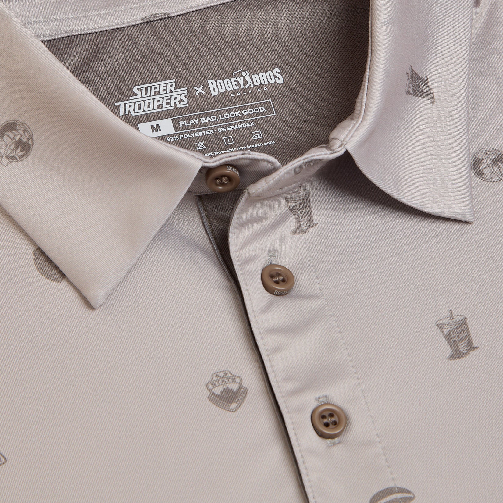 Close-up of a beige collared shirt with Super Troopers x Bogey Bros branding on the tag. The shirt features brown buttons and small printed graphics of drinks and badges. The fabric is 91% polyester, 9% spandex.