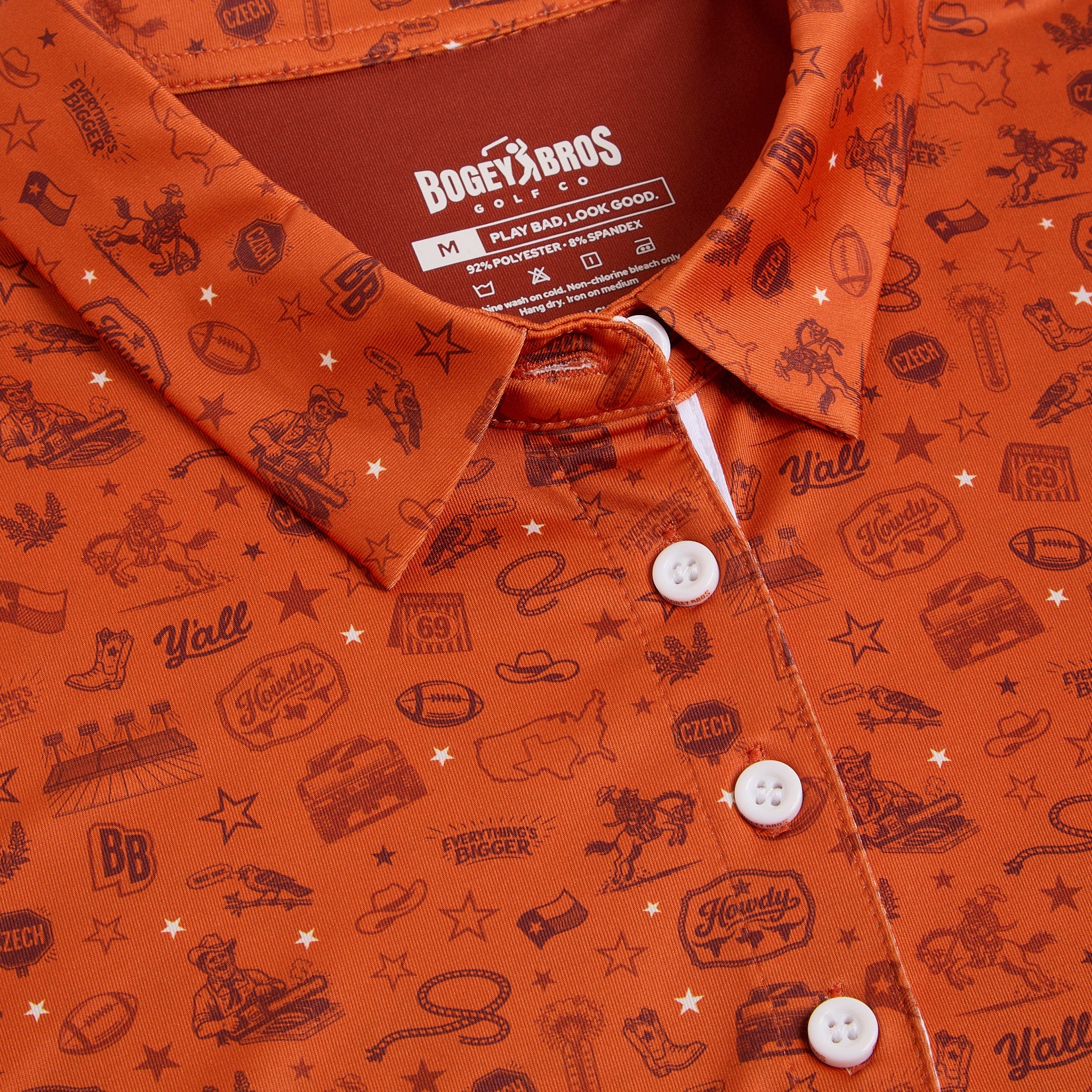 Close-up of an orange collared shirt with white buttons, featuring playful western-themed graphics like cowboys, cowboy boots, stars, footballs, and words such as “Y’all,” “Howdy,” and “Everything’s Bigger.” Shirt tag reads “BOGEY BROS CLUB.”.