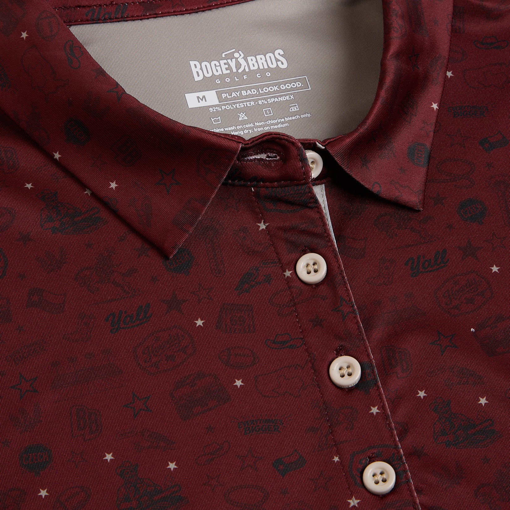 Close-up of a maroon polo shirt with four beige buttons, featuring various small, dark patterns and playful graphics, including stars, icons, and the word Yall. The inside label reads Bogey Bros and provides size and fabric information.