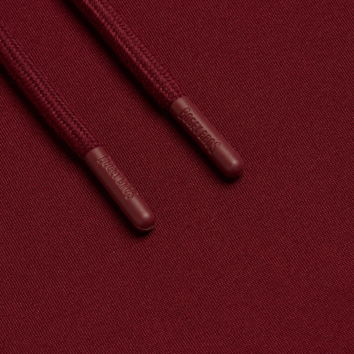 Close-up of two burgundy drawstrings from the Burgundy - Hoodie, featuring textured aglets embossed with the word DREW HOUSE. The matching burgundy fabric has a fine knit texture and is known for its moisture-wicking properties, shown here under even, neutral lighting.