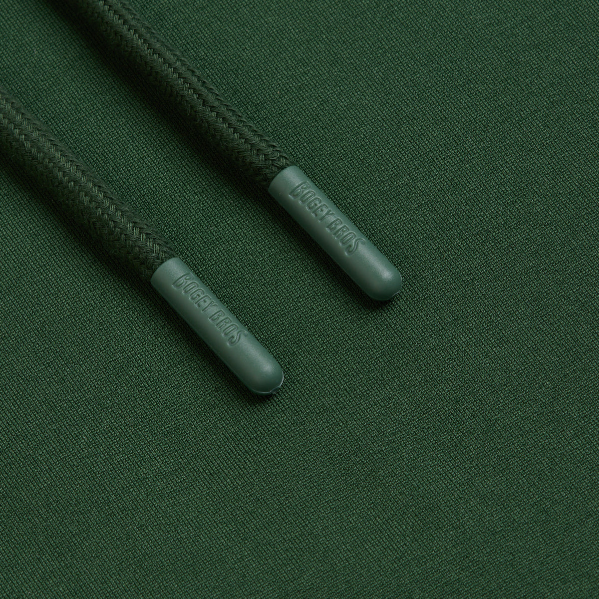Close-up of two dark green drawstrings from the Forest Green - Hoodie resting parallel on matching forest green fabric. The plastic aglets are embossed with LAGRE JEANS and positioned near the top left atop a smooth, tightly woven textile background similar to moisture-wicking athletic shorts.