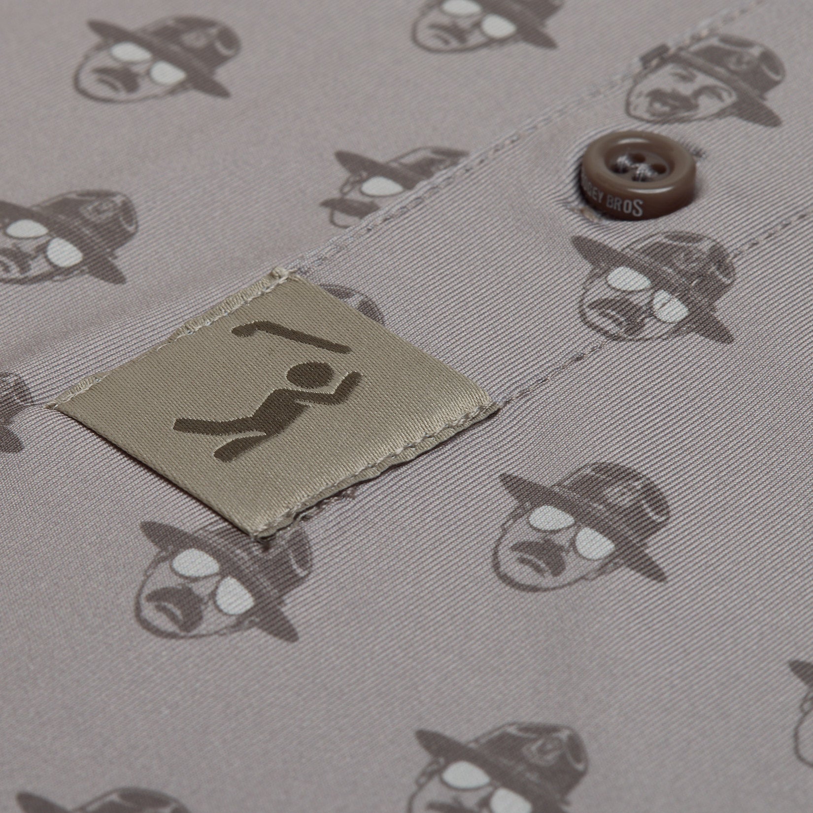 Close-up of taupe fabric with repeated dark print of a hat-and-glasses face, a stitched square patch showing a simplified person with arms up, and two brown buttons partially visible near the top right. The overall color palette is muted.
