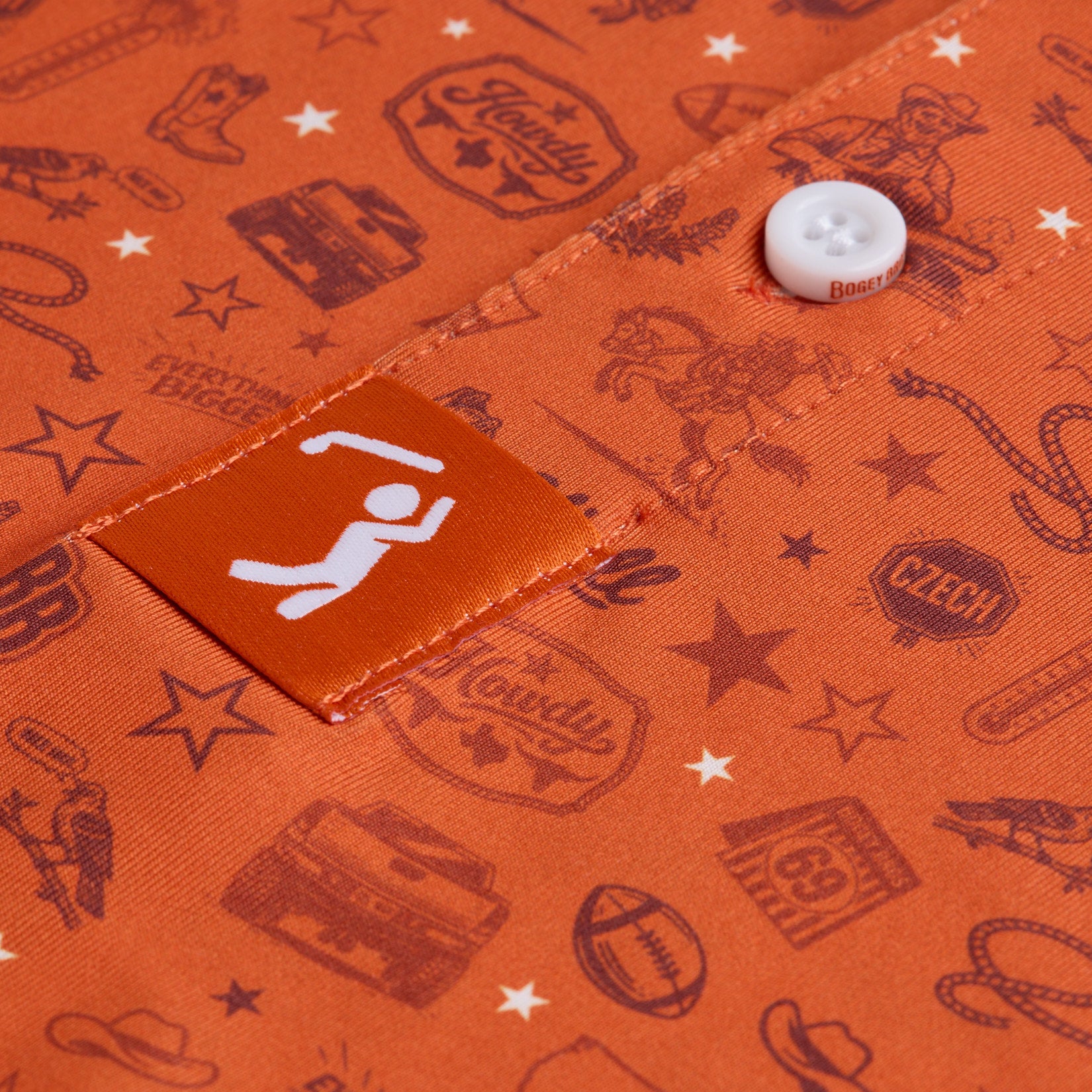 Close-up of an orange shirt with white buttons, featuring western-themed graphics like cowboys, boots, stars, ropes, truck, and signs. An orange label with a white swinging figure is stitched near the button. The fabric pattern uses dark red-brown ink.
