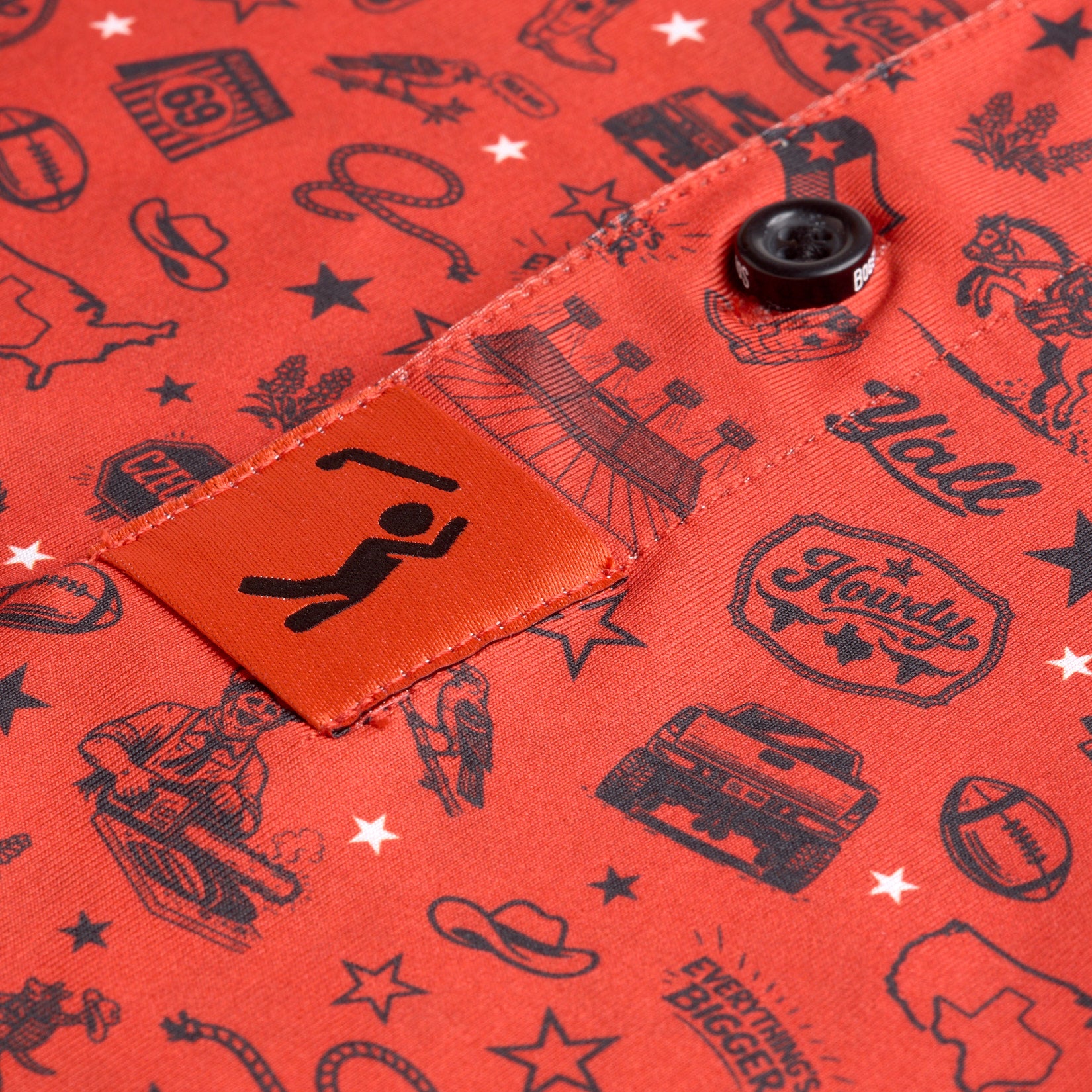 Close-up of red fabric with black Western-themed illustrations, including cowboy hats, boots, trucks, footballs, and stars. A tag with a stylized person raising arms is sewn near a black button. Stitching and fabric texture are visible throughout.