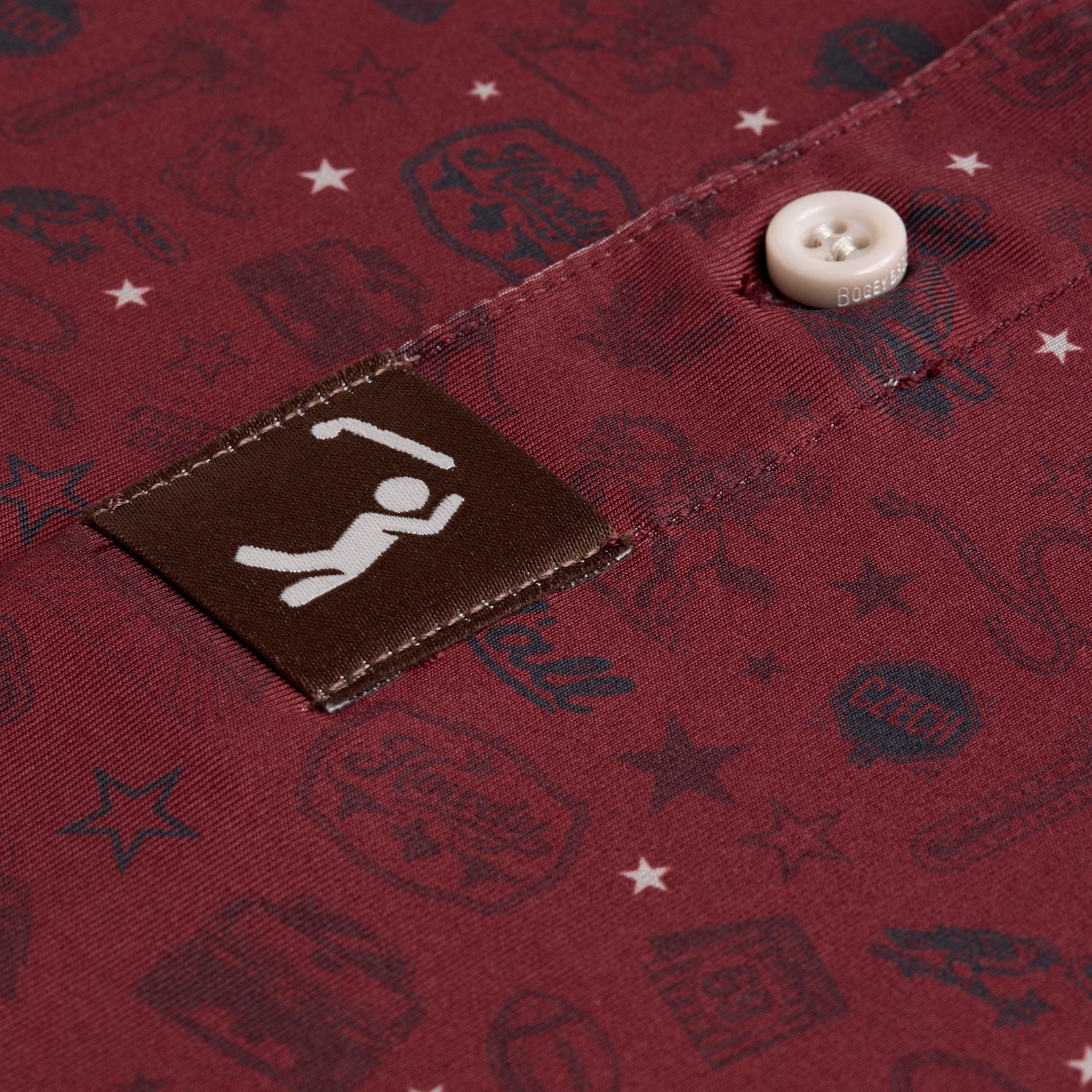Close-up of a maroon shirt with dark blue printed motifs, a white button, and a brown fabric tag. The tag shows a white icon of a person lifting a barbell. Pattern elements include stars, labels, and abstract designs on the fabric.