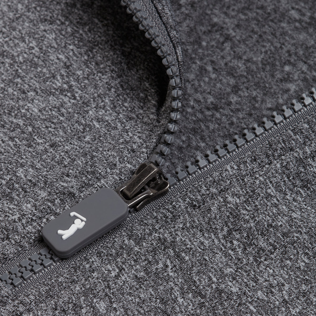 A close-up of the Heather Gray - Q-Zip shows a dark gray, rectangular matte zipper pull with a small white logo on textured, heather gray moisture wicking fabric. The athletic fit garment features partially open zipper teeth and visible stitching.