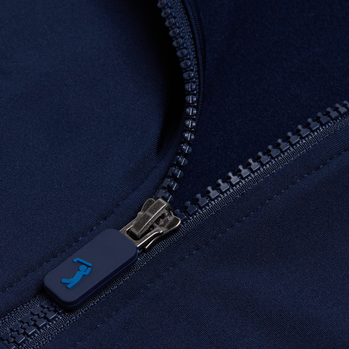 A detailed close-up of the Navy - Q-Zip highlights its athletic fit, featuring a matte blue zipper pull adorned with a small blue logo resembling a person stretching. The neatly stitched zipper teeth and surrounding dark blue fabric showcase the product’s high-quality texture and craftsmanship.