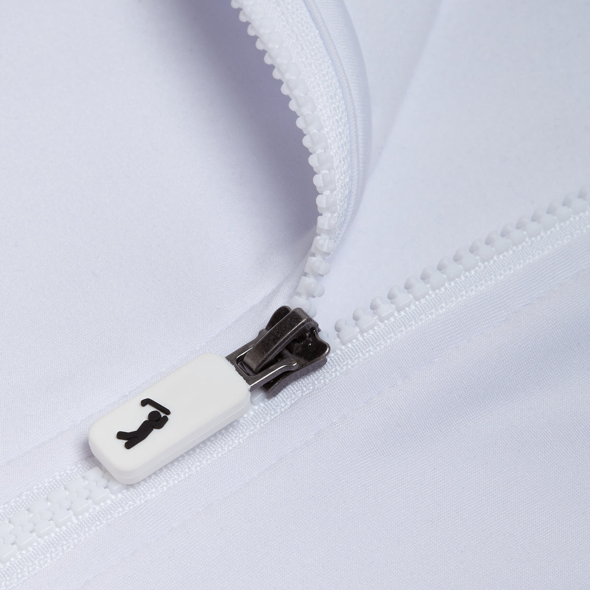Close-up of the White - Q-Zip’s white zipper, partially unzipped against matching white fabric. The zipper pull is white, featuring a black icon resembling a person. The visible zipper teeth and stitching highlight the garment’s texture and material details. The fabric also provides UPF 50 protection for enhanced sun safety.