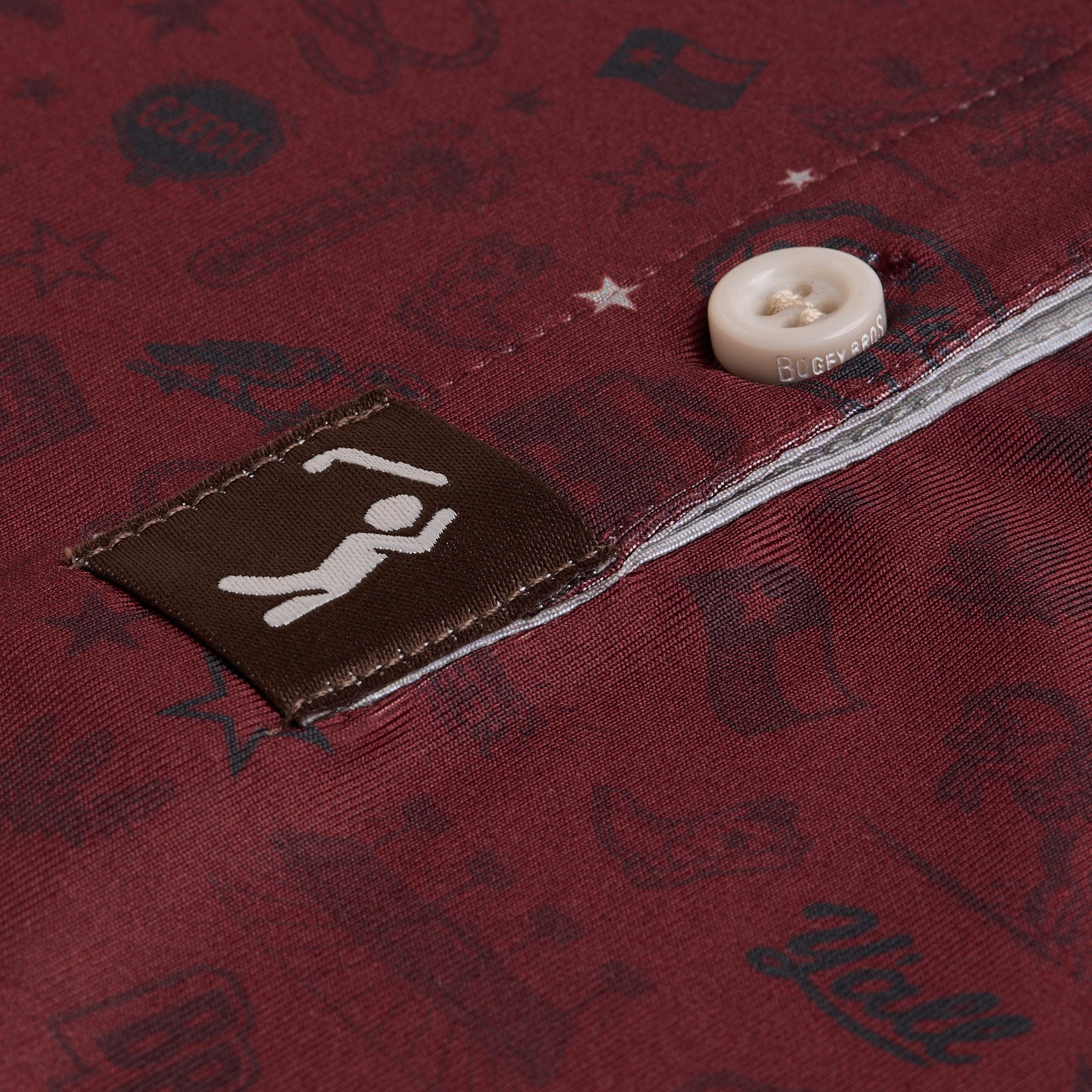 Close-up of a maroon fabric with faint, dark illustrations and a white button. A brown tag with a simple white stick figure lifting weights is sewn onto the fabric near the button. White stitching runs along the edge of the button placket.