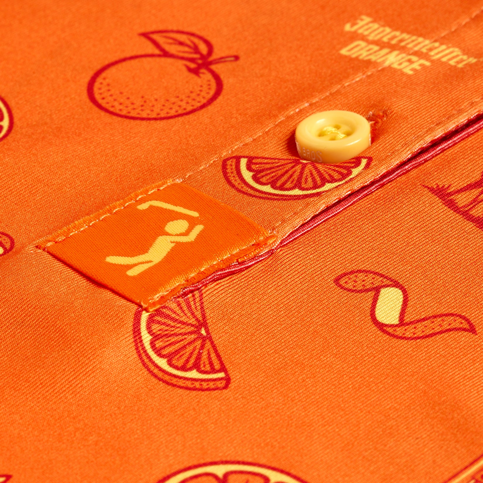 Close-up of the Main Squeeze - Women's Polo: an orange shirt with a yellow button and fruit print, featuring a small white icon of a person lifting a barbell near the seam. The smooth, glossy fabric offers moisture-wicking comfort.
