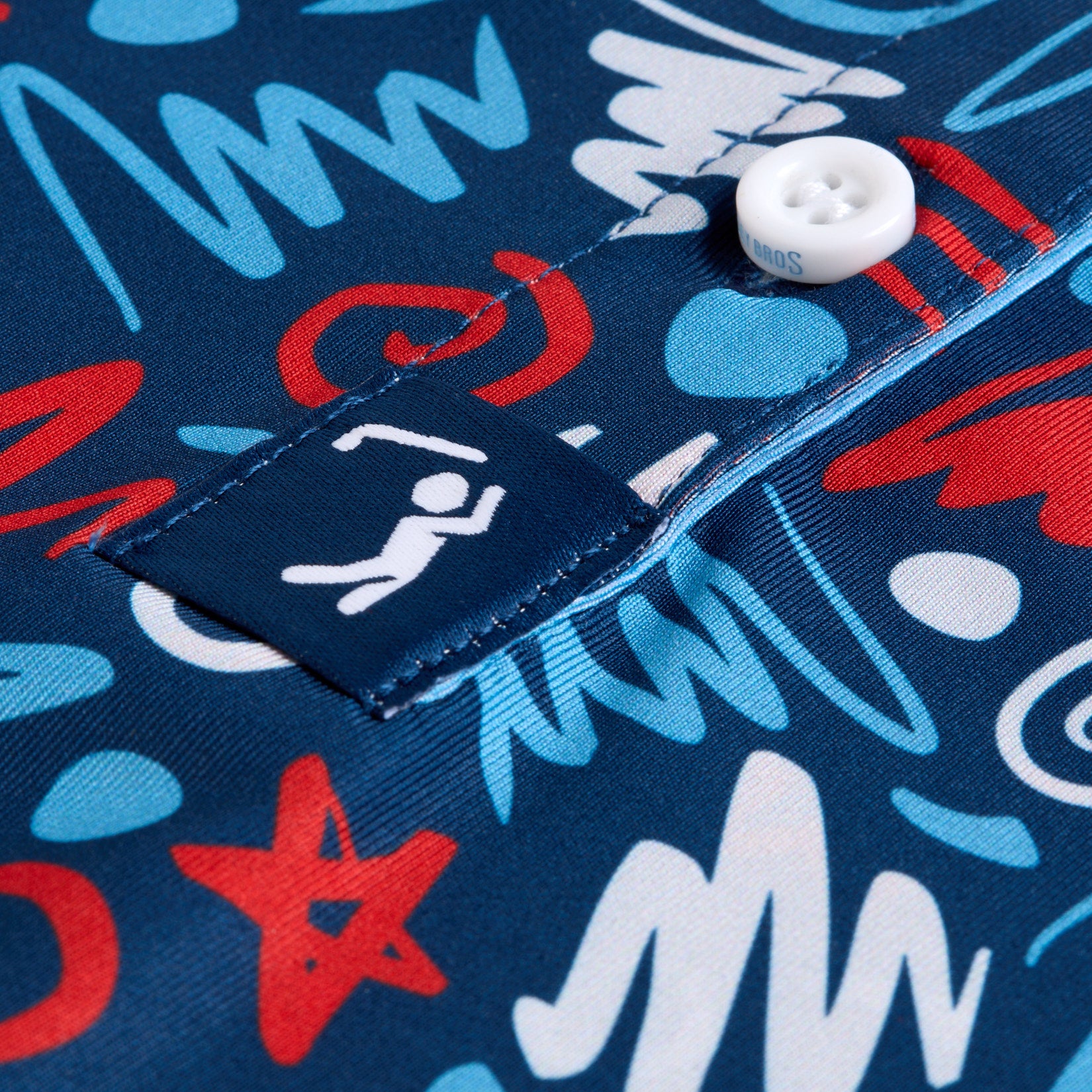 Close-up of a blue shirt with a colorful abstract red, white, and blue pattern. A white button is sewn onto a placket, and a navy tag with a white icon of a person lying under a roof is stitched on the edge of the placket.