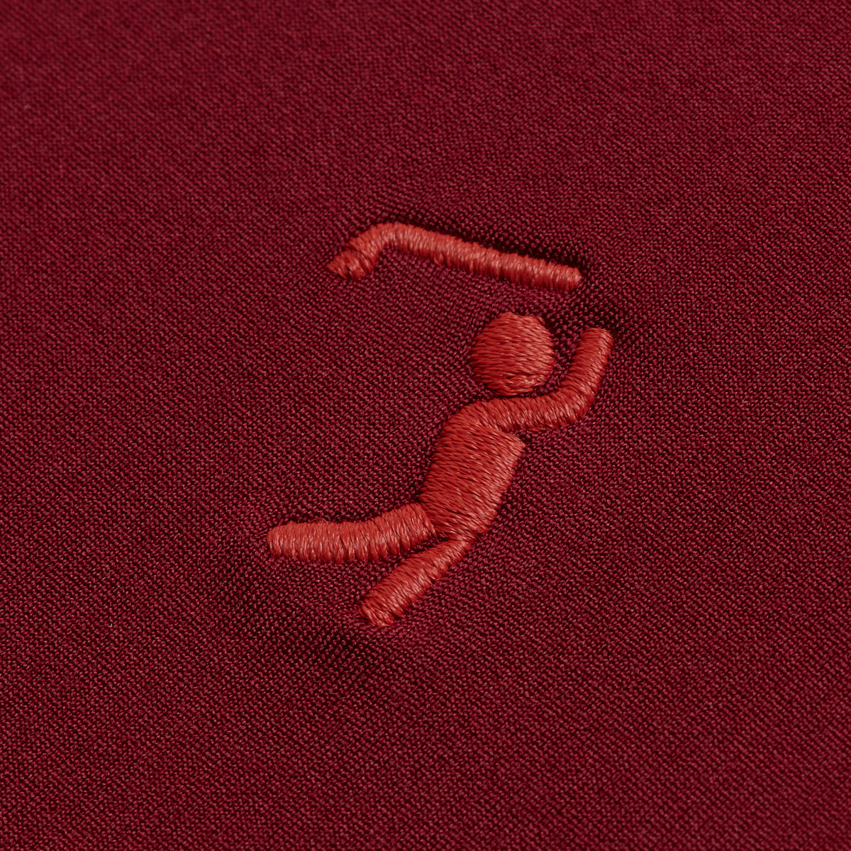 A red embroidered figure on burgundy fabric appears to jump or dance with one arm raised and bent, and one leg lifted. The simple, stylized design is perfect for the Burgundy Hoodie, offering both style and comfort.