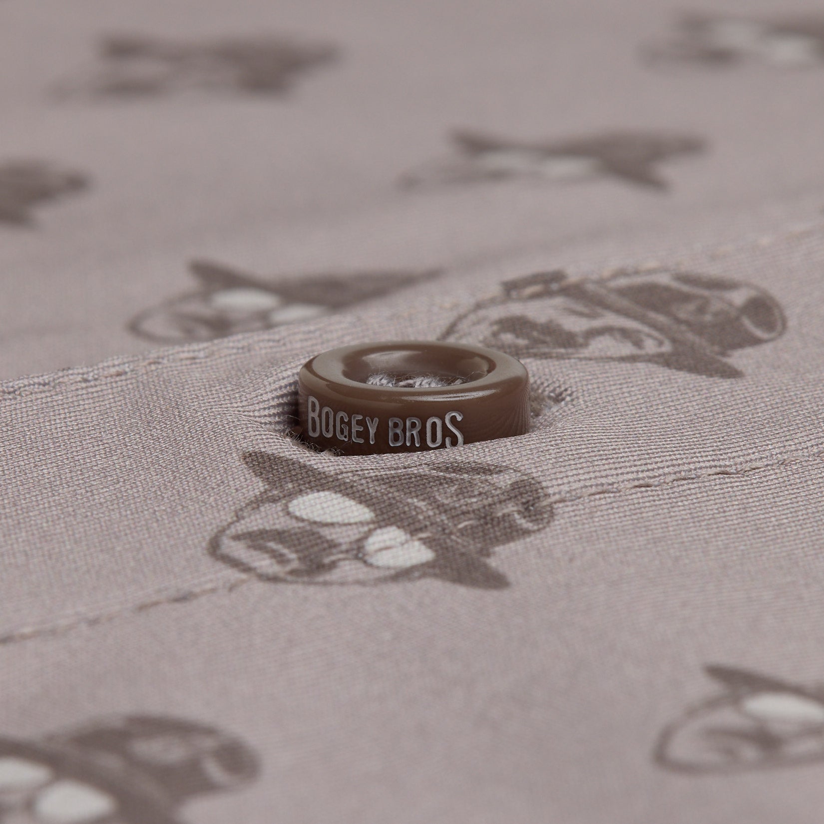 A brown button reading BOGEY BROS is sewn onto light brown fabric printed with repeating cartoon faces in sunglasses and hats. The button sits along a stitched seam, blending with the illustrated pattern in the background.