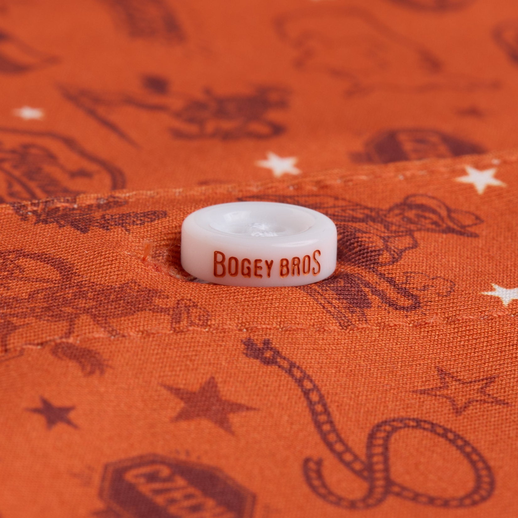A white button with Bogey Bros in orange text is sewn onto orange fabric printed with brown western-themed images, including stars, a cowboy rope, and various cowboy illustrations. The button is centered in the image.