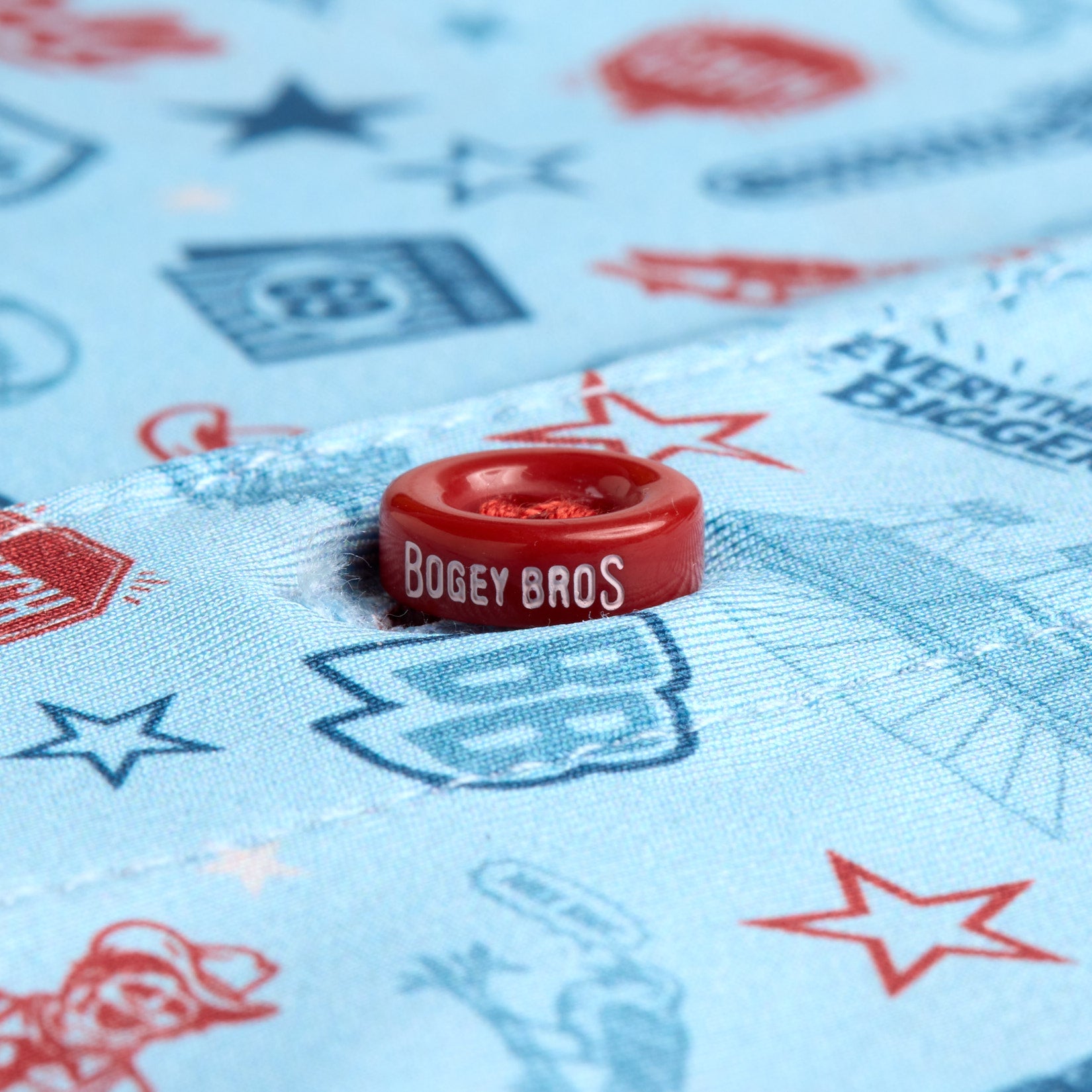 A close-up of a red button with BOGEY BROS printed on it, attached to light blue fabric featuring playful red and blue illustrations and patterns including stars, shapes, and generic logos.