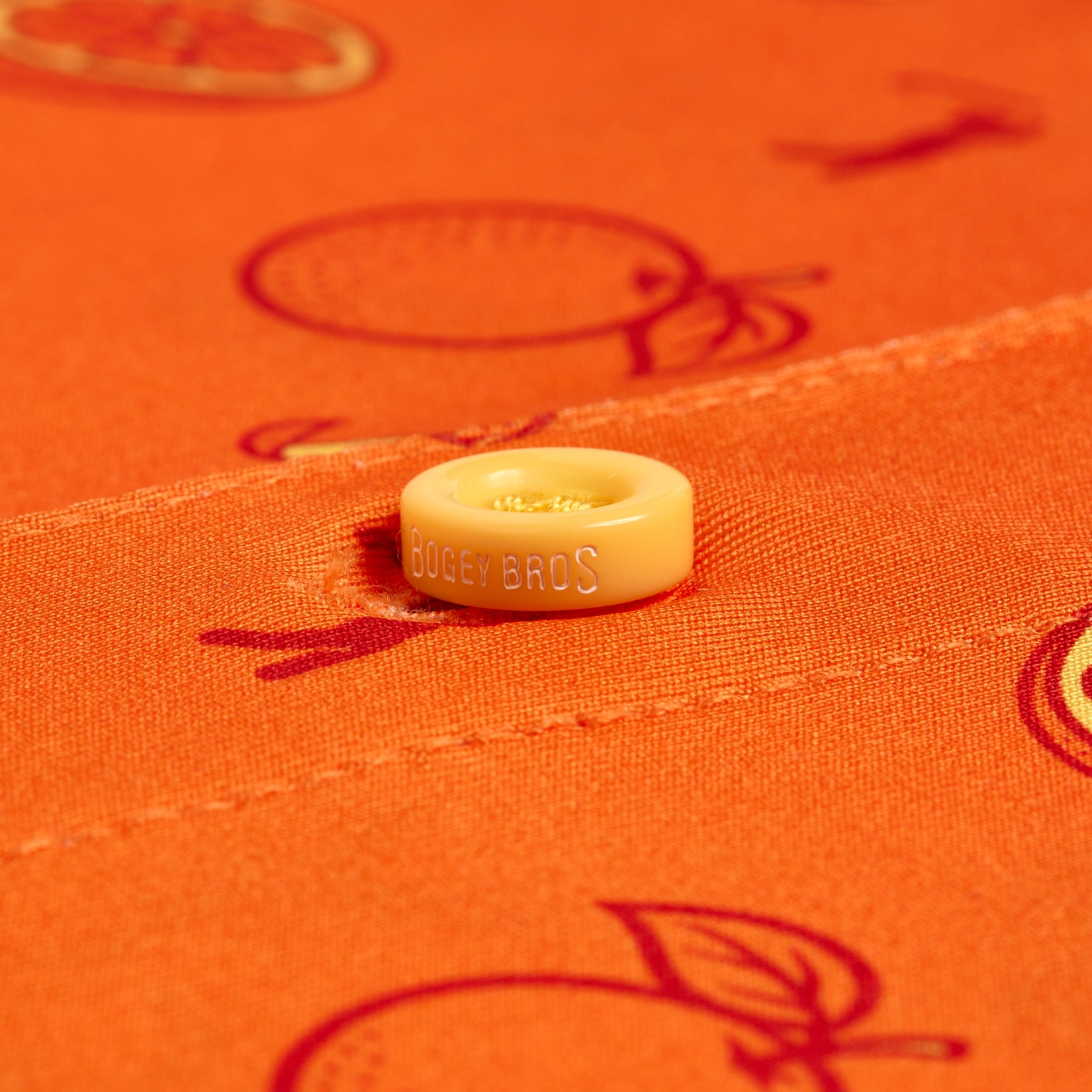 A close-up of the Main Squeeze - Polo shows an engraved orange BOGEY BROS button sewn onto moisture-wicking, bright orange fabric patterned with red-outlined oranges and leaves, emphasizing its citrus-inspired design.