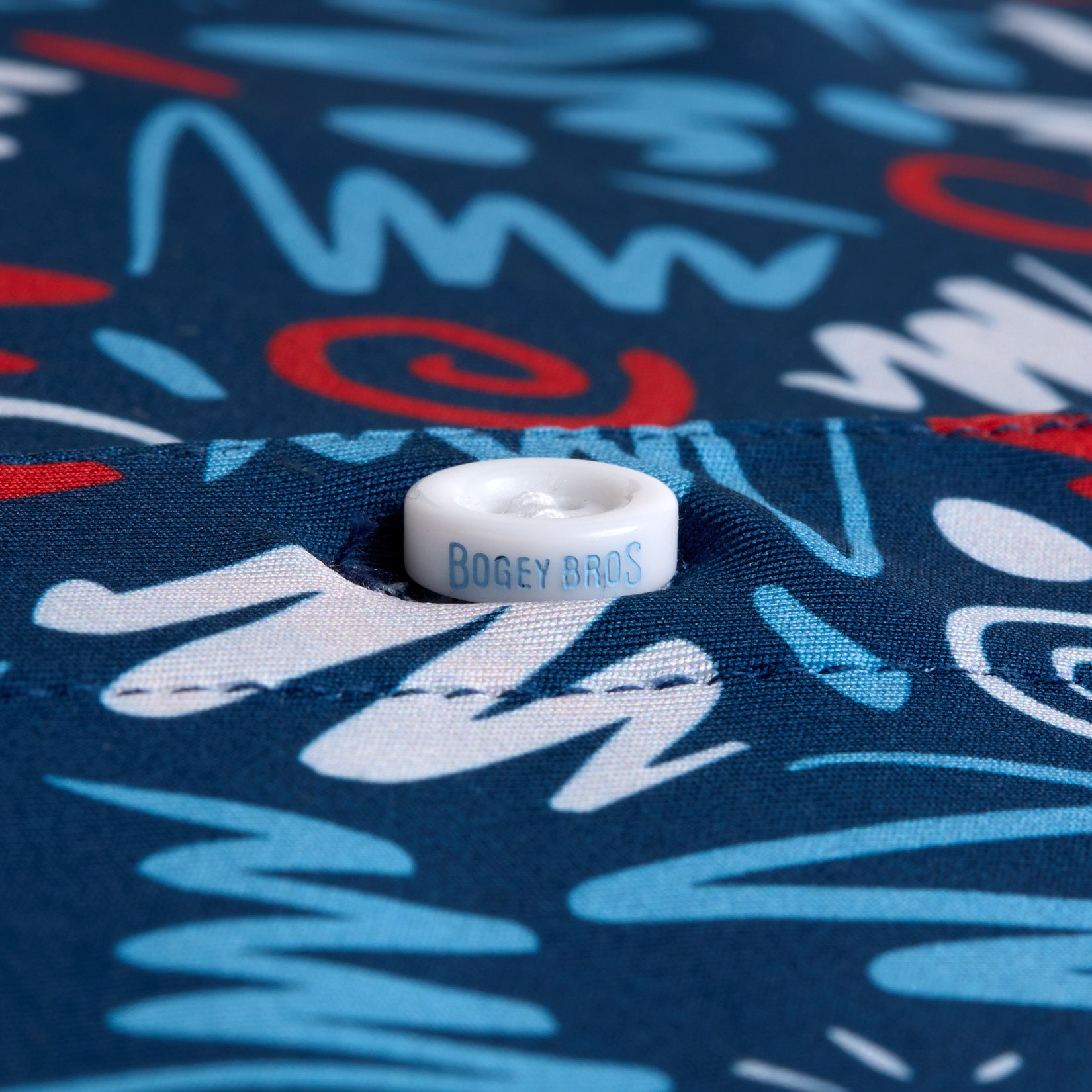 Close-up of a white button sewn onto fabric with BOGEY BROS printed on it in blue. The fabric features an abstract pattern of white, blue, and red squiggles and lines on a navy blue background.
