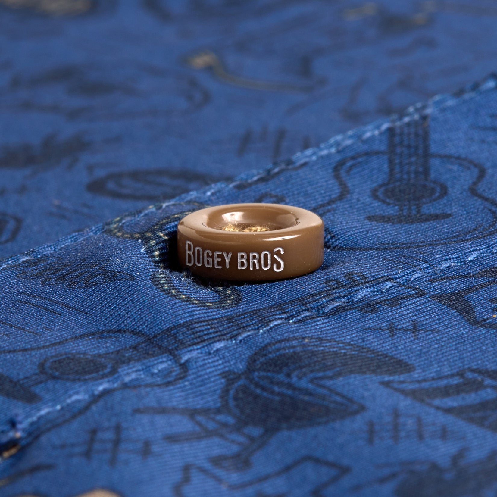 Close-up of a brown button with "BOGEY BROS" in white text on the Ringling Road - Polo, featuring blue fabric patterned with dark icons like guitars and golf clubs. Blue stitching runs diagonally across the fabric beneath the button.