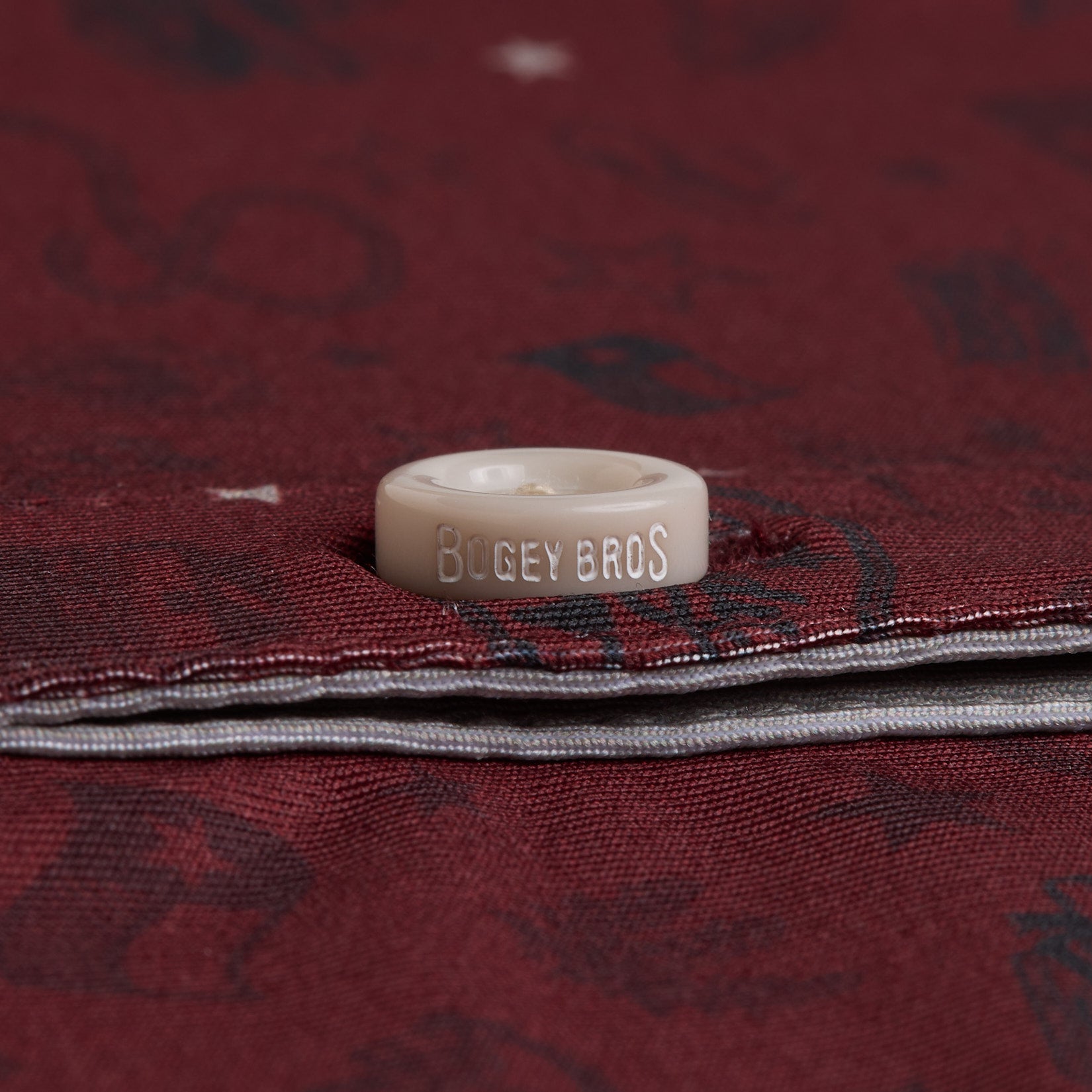 A close-up of a beige button engraved with BOGEY BROS sewn onto dark red, patterned fabric. The button is attached near the edge where two layers of fabric overlap, with some white contrast stitching visible.