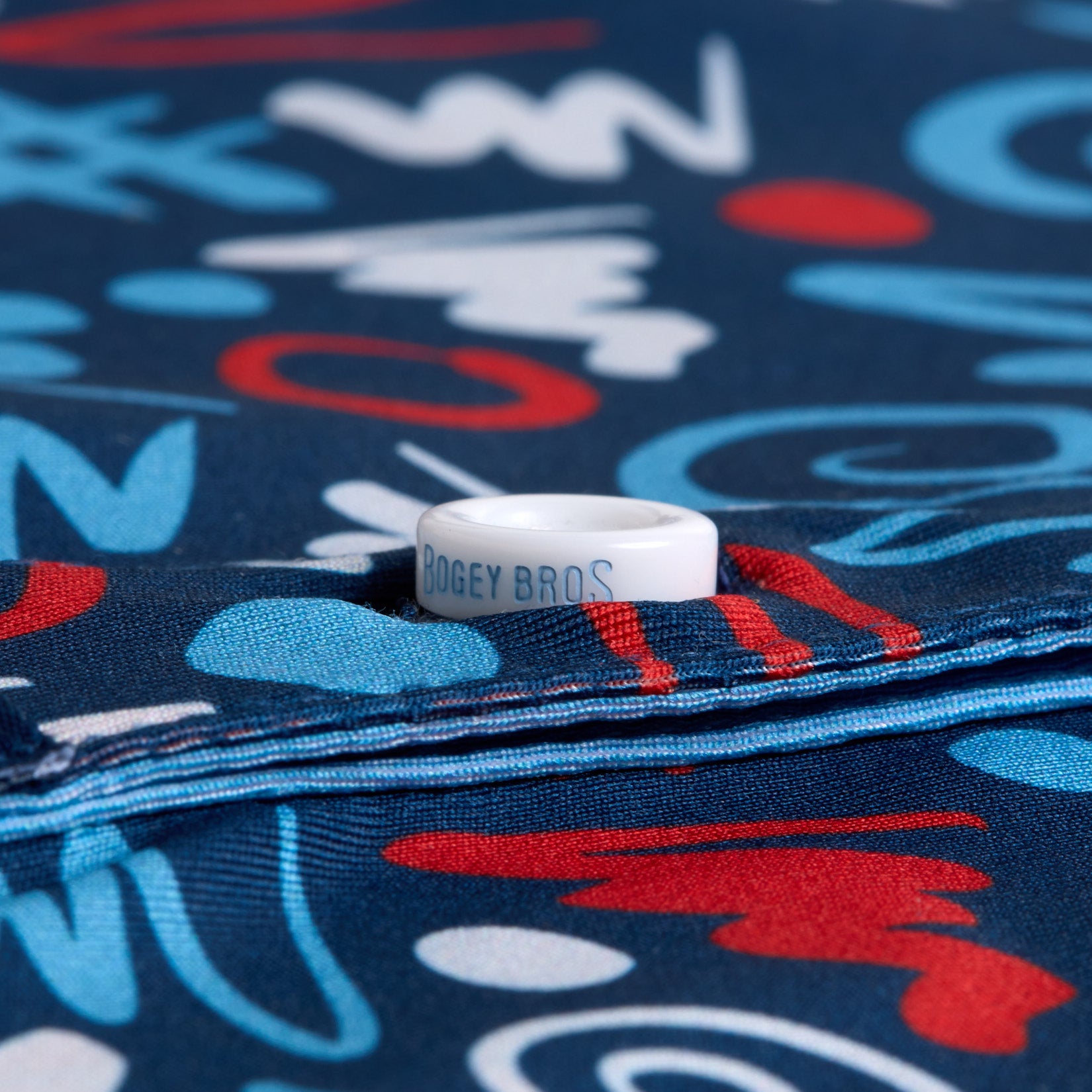 A white ring with “BOGEY BROS” printed on it rests on folded blue fabric with an abstract red, white, and blue pattern of scribbles and shapes. The focus is on the ring’s text and the colorful, layered fabric beneath it.