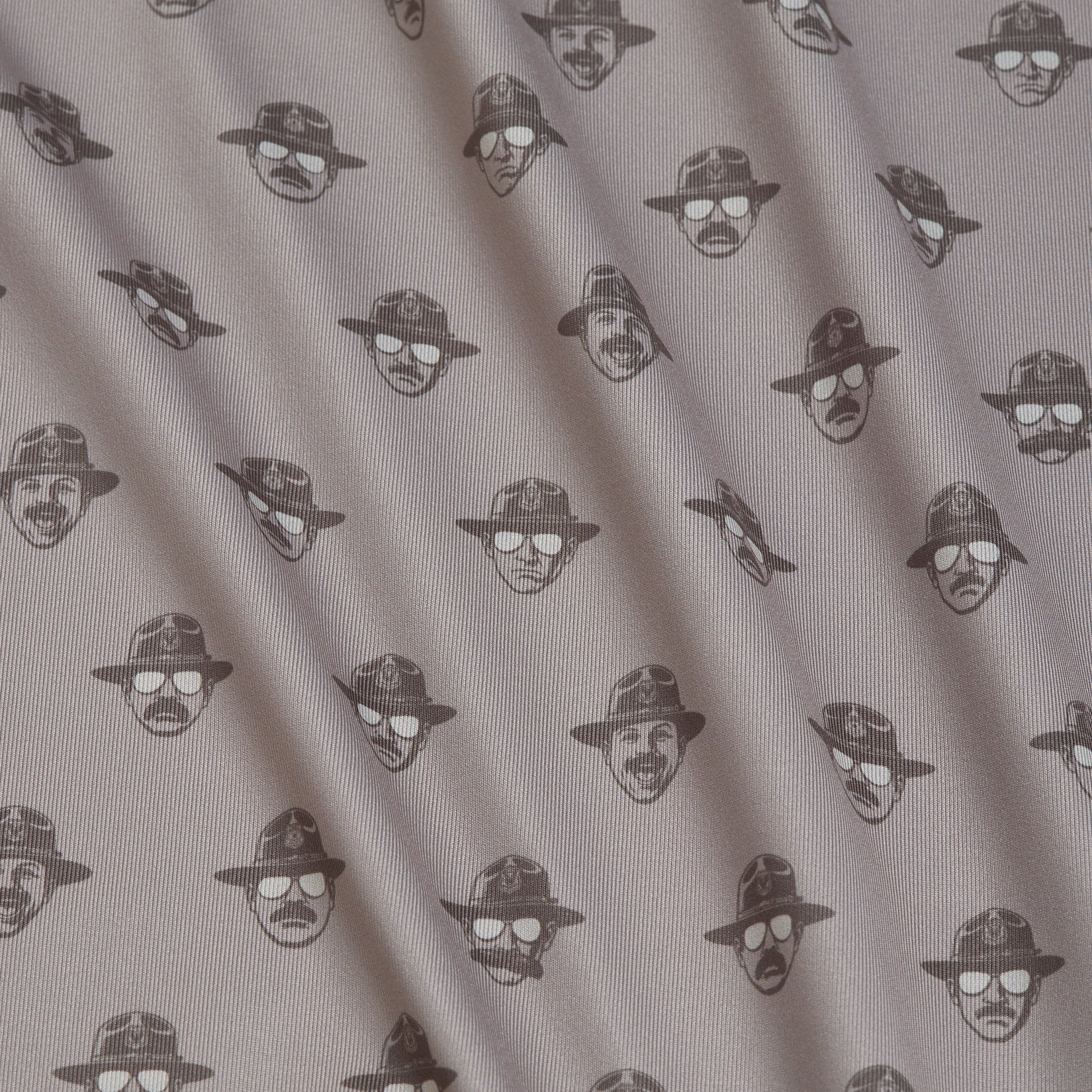 Patterned fabric with small, repeating black line drawings of faces wearing hats and sunglasses on a light background. The fabric has soft folds and diagonal shadows, giving it a textured look. Each face is spaced evenly in rows.