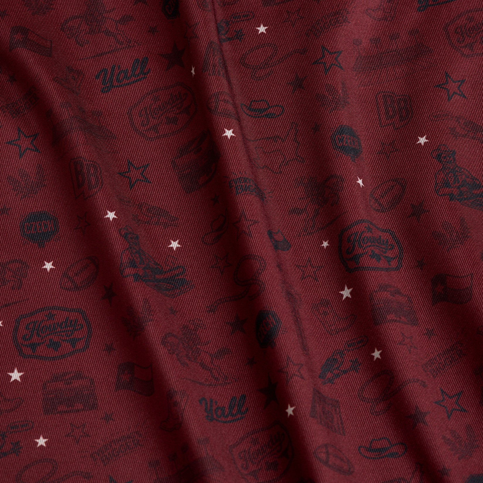 Maroon fabric with small white stars and dark Western-themed icons, including cowboy hats, boots, boots, horseshoes, “Y’all” text, a sheriffs badge, guitars, and other rodeo motifs, scattered throughout. The fabric has a slight textured, wavy appearance.