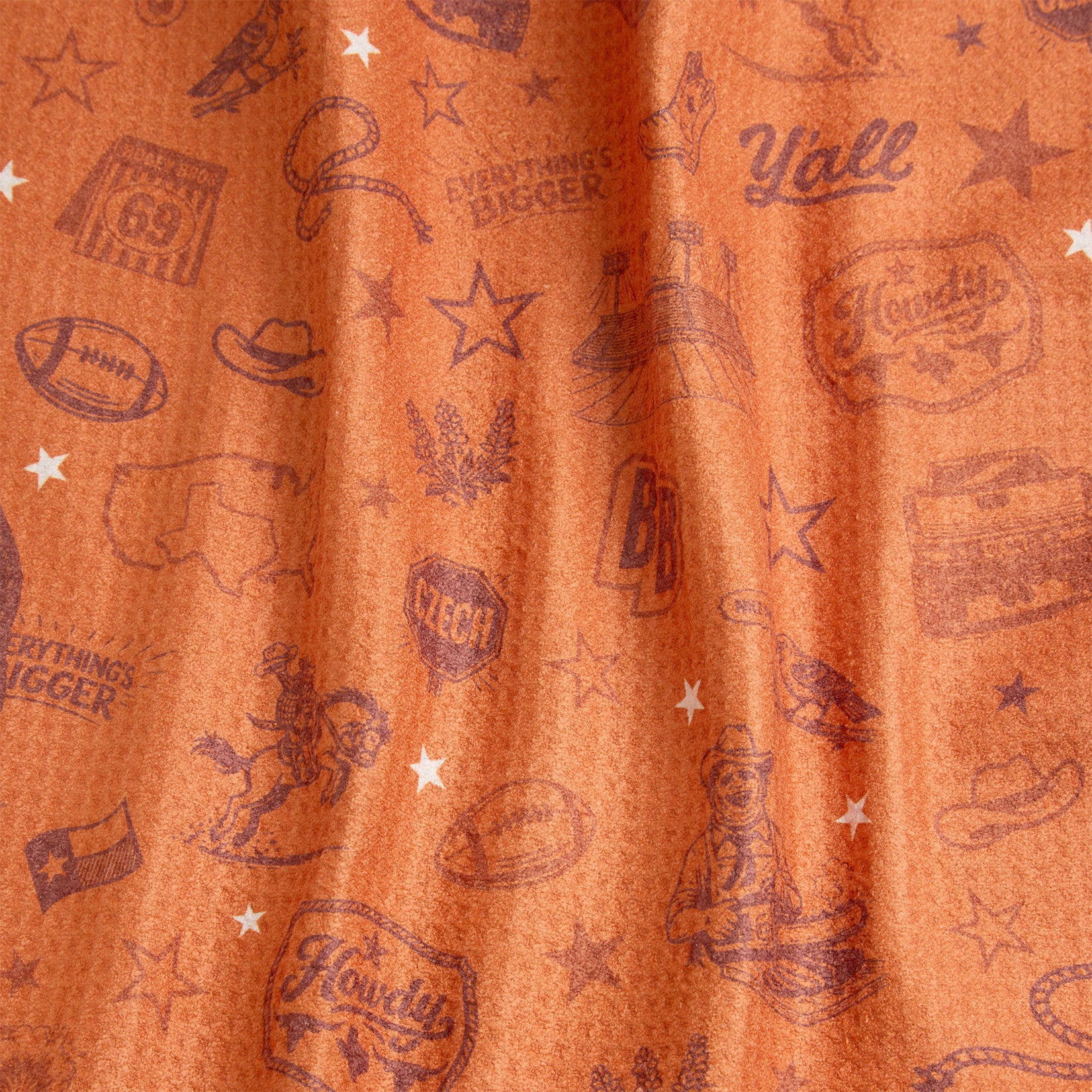 Orange fabric with western-themed illustrations, including cowboy hats, boots, stars, a map of Texas, horseshoes, footballs, and phrases like Howdy, Yall, and Everything’s Bigger. White stars are scattered throughout the pattern.