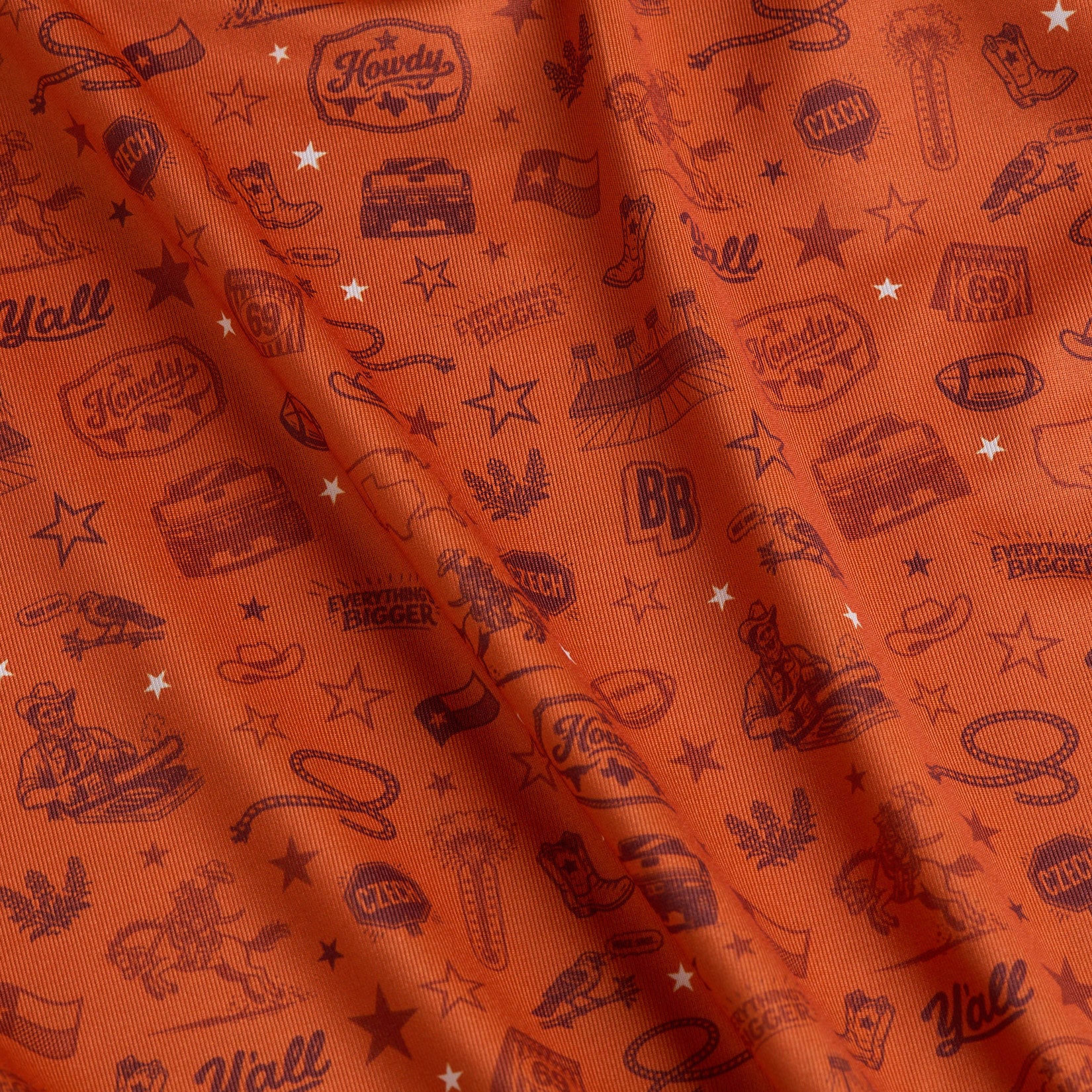 Orange fabric with repeating maroon Texas-themed illustrations, including cowboy hats, boots, armadillos, stars, footballs, BBQ grills, cacti, and words like “Howdy,” “Y’all,” and “Everything’s Bigger.” The fabric is lightly wrinkled.
