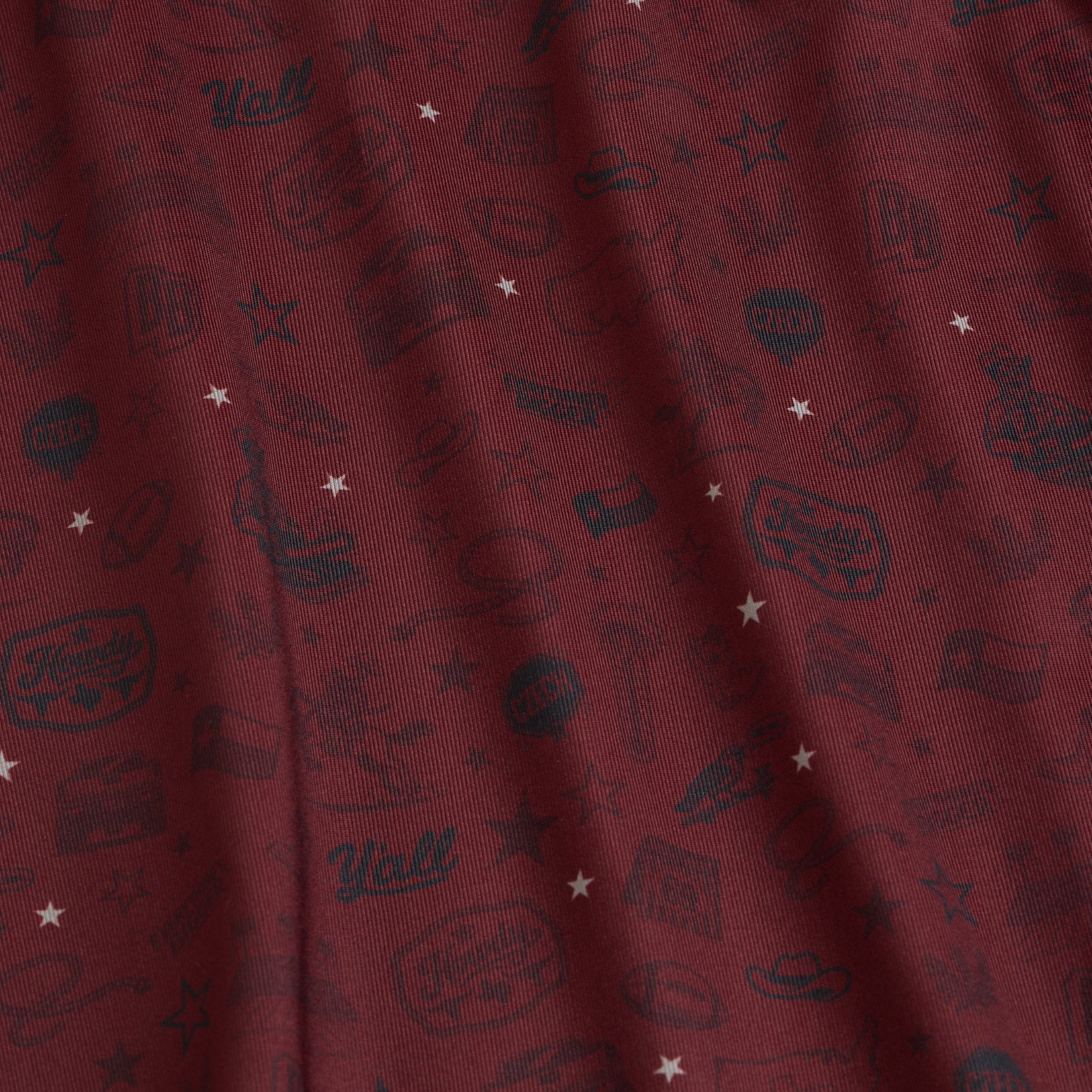 Close-up of maroon fabric with small black sketches of cowboy hats, boots, and bandanas, mixed with white stars and Yall! text. The surface is slightly wrinkled, making the designs appear at varying angles and depths.