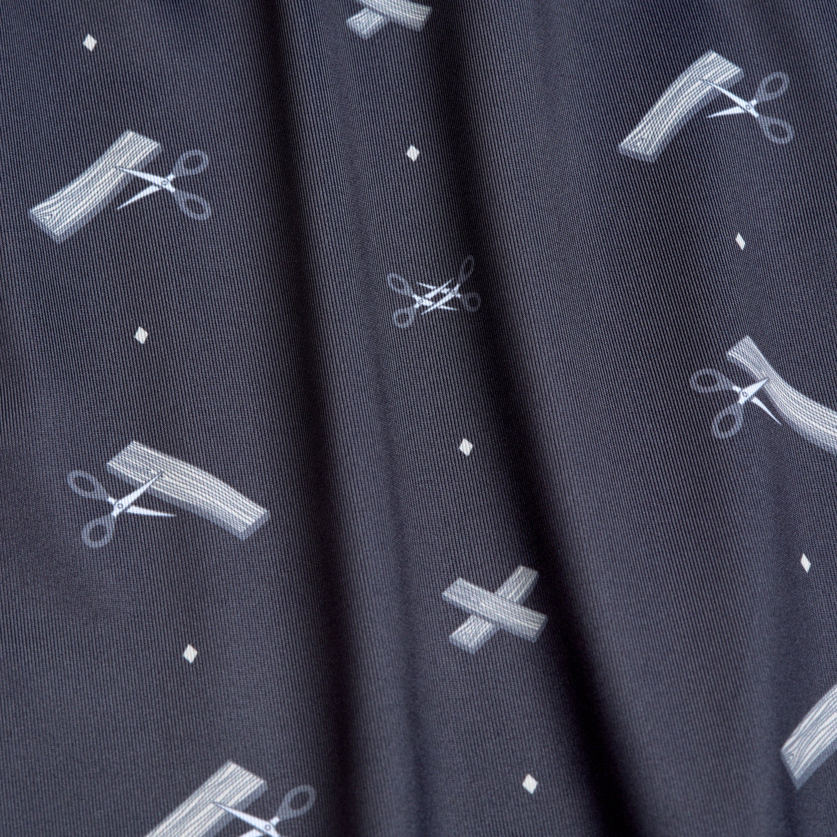 A close-up of the Scissor Me Timbers - Women's Polo shows dark, moisture-wicking fabric with white scissors, cut lines, Xs, and diamonds in a whimsical pattern; athletic fit fabric displays texture and a slight vertical fold.